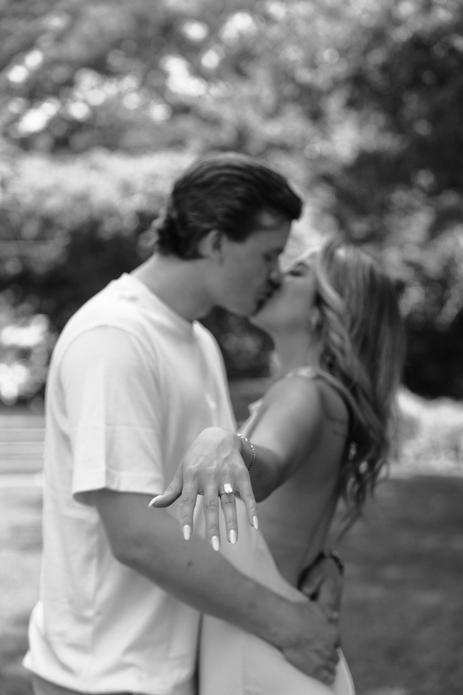 Romantic black and white photo of a couple sharing a kiss outdoors while showing off an engagement ring.