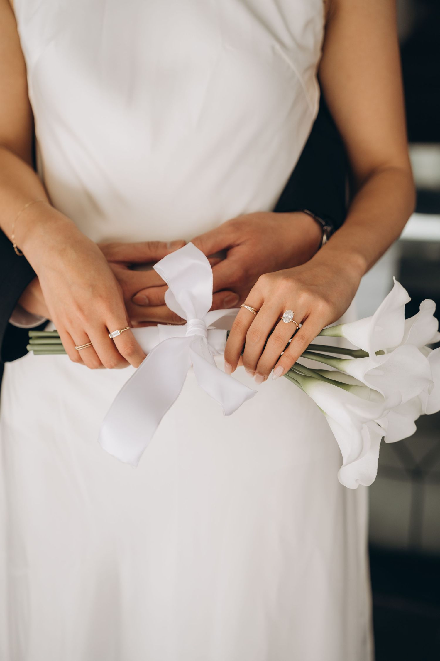 Close-up of hands in white wedding dress holding white ribbon and lilies during bridal preparation.