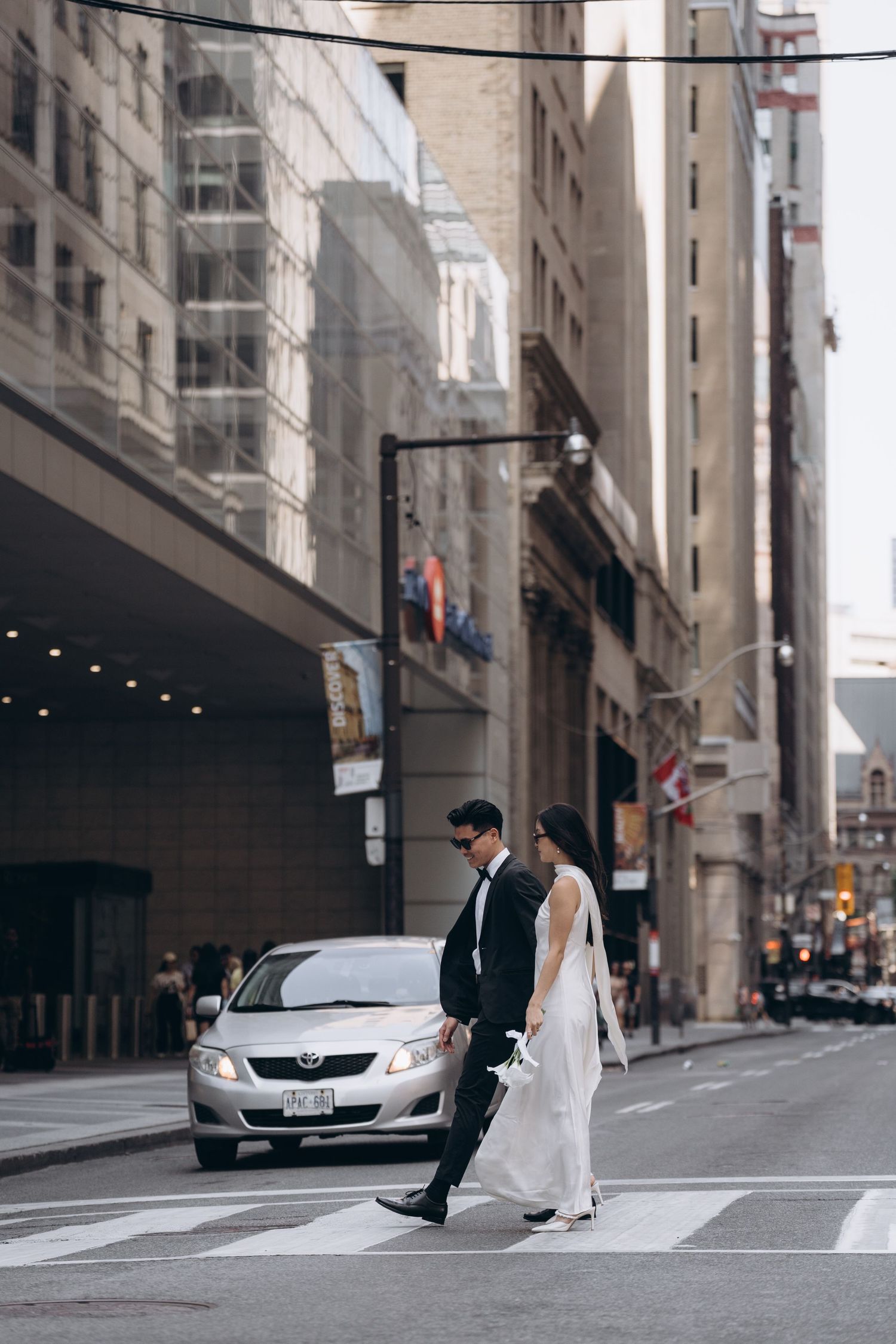 A couple in wedding attire crosses a downtown street with tall buildings and a silver car in the background.