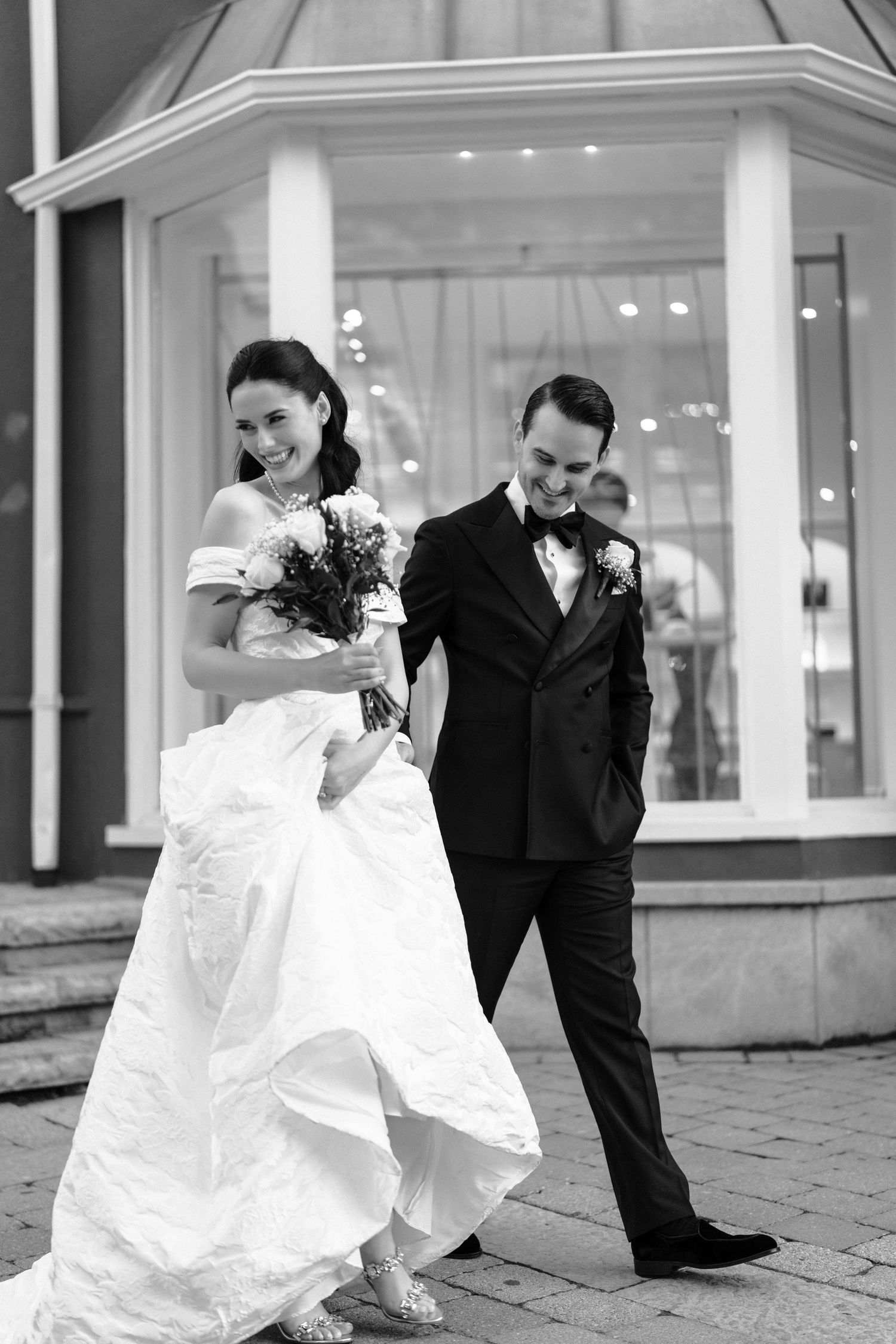 A black and white wedding photo of a couple in formal attire standing outside an elegant building entrance.