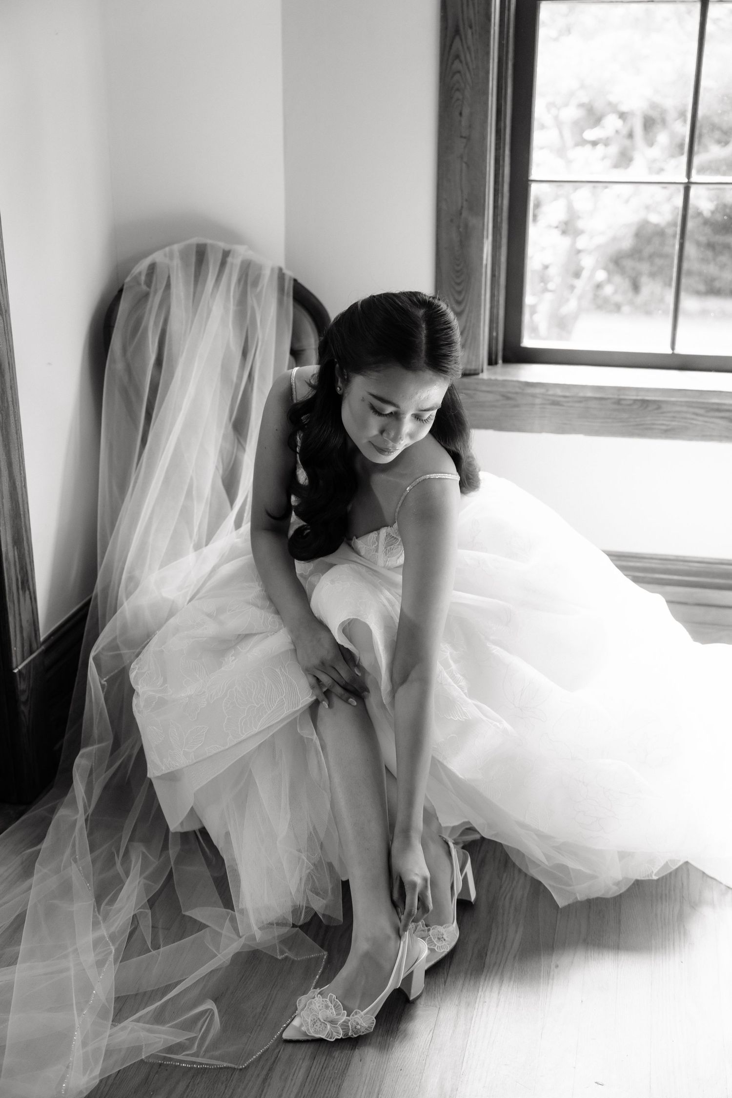 A series of black and white photos showing a bride in a flowing white wedding gown adjusting her shoes by a window.