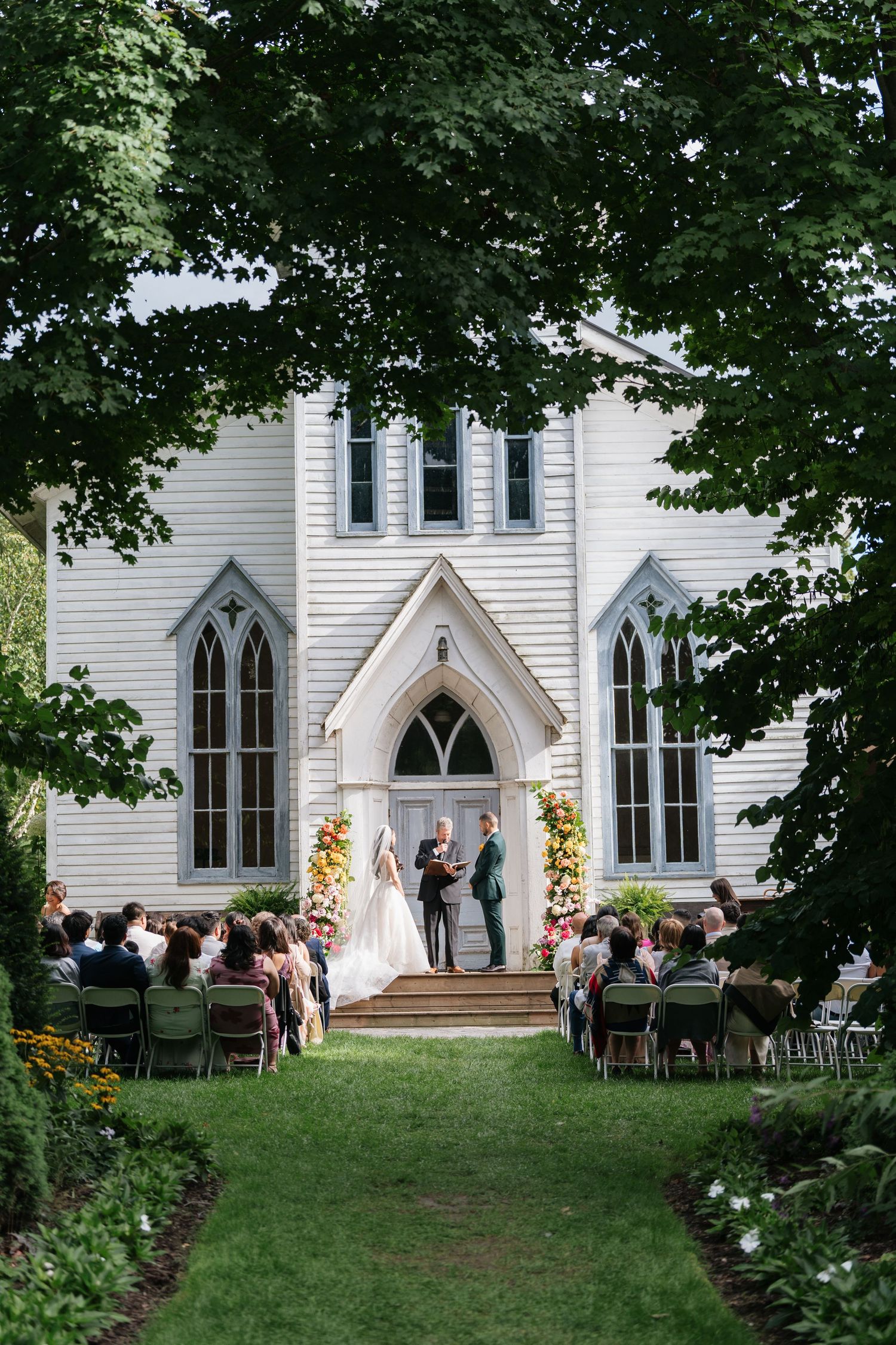 A wedding ceremony takes place at a charming white Gothic-style chapel with pointed arched windows and floral arrangements.