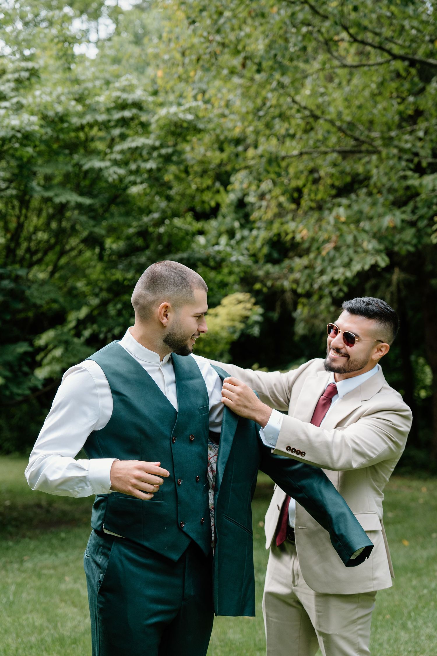 Two men in formal attire help each other get dressed outdoors, with one adjusting the other's green vest in a garden setting.