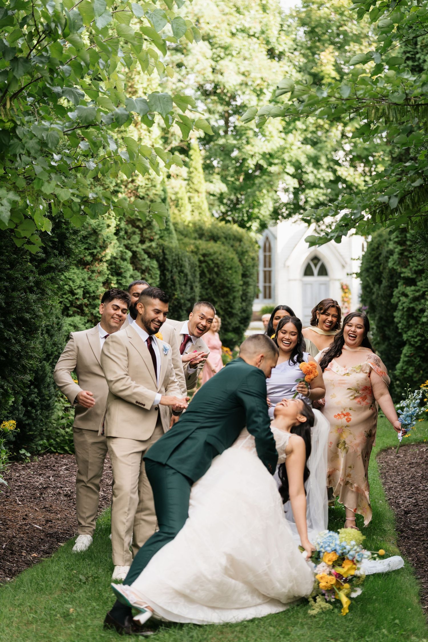 Wedding party poses playfully in a garden setting with bridesmaids in tan dresses and groomsmen in matching suits.