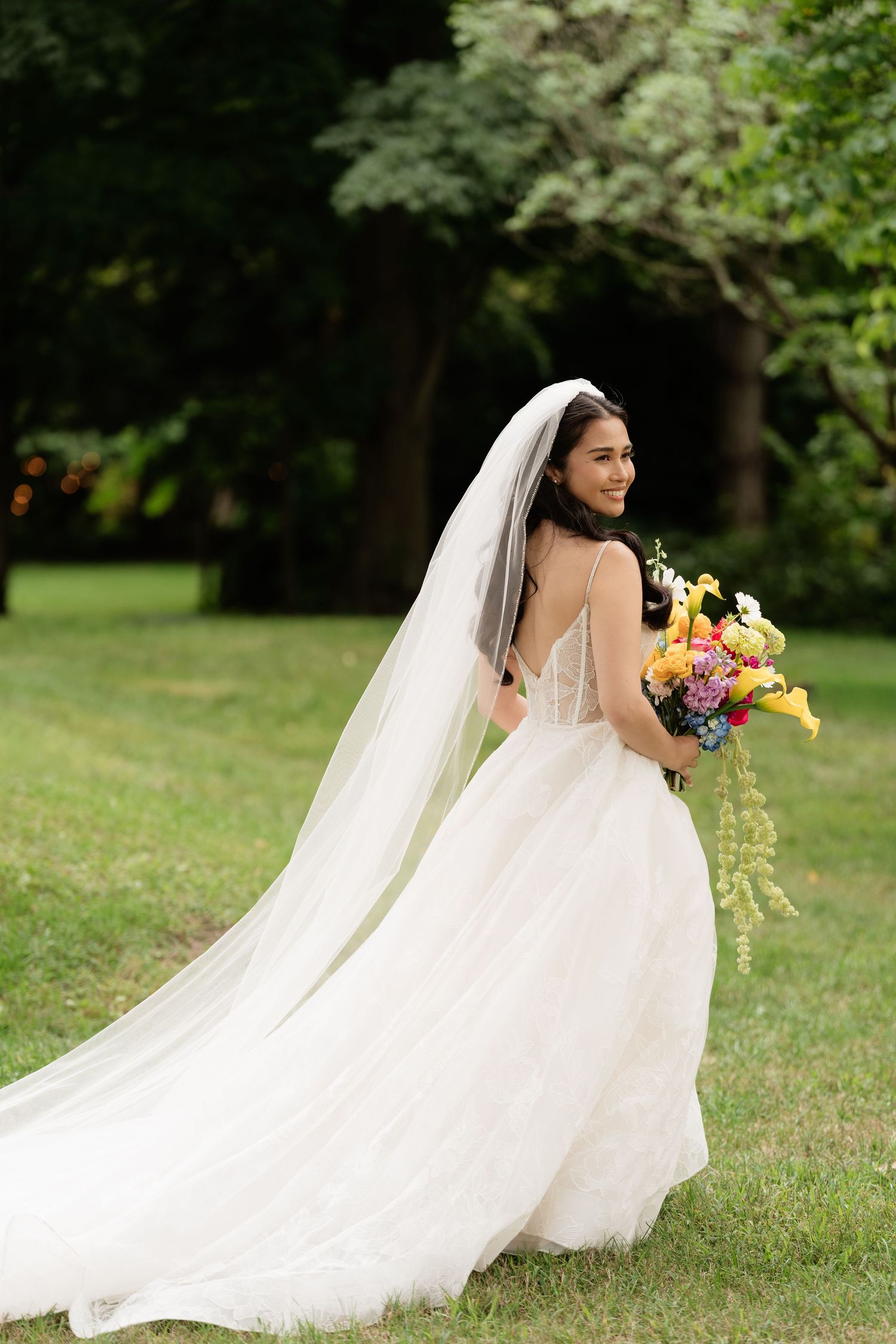 Bride in flowing white dress walks through garden holding colorful bouquet with trailing veil.