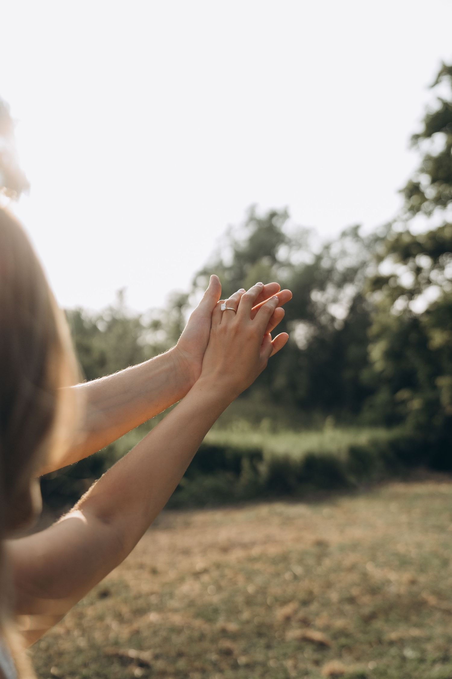 Hands raised in prayer against a sunlit natural outdoor background with trees and grass.