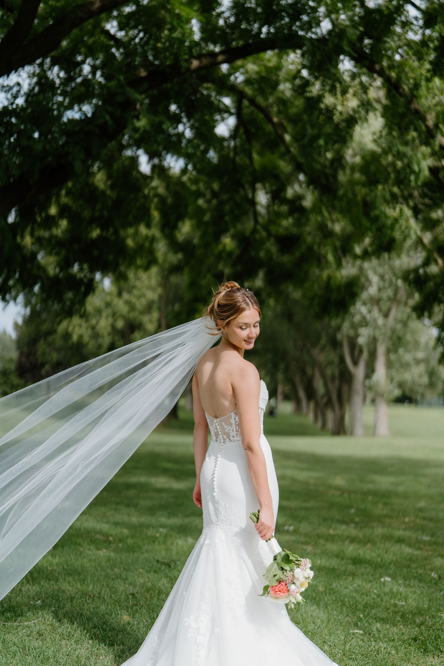 A flowing white wedding veil catches the breeze in a lush green garden setting as a bride poses in her lace gown.