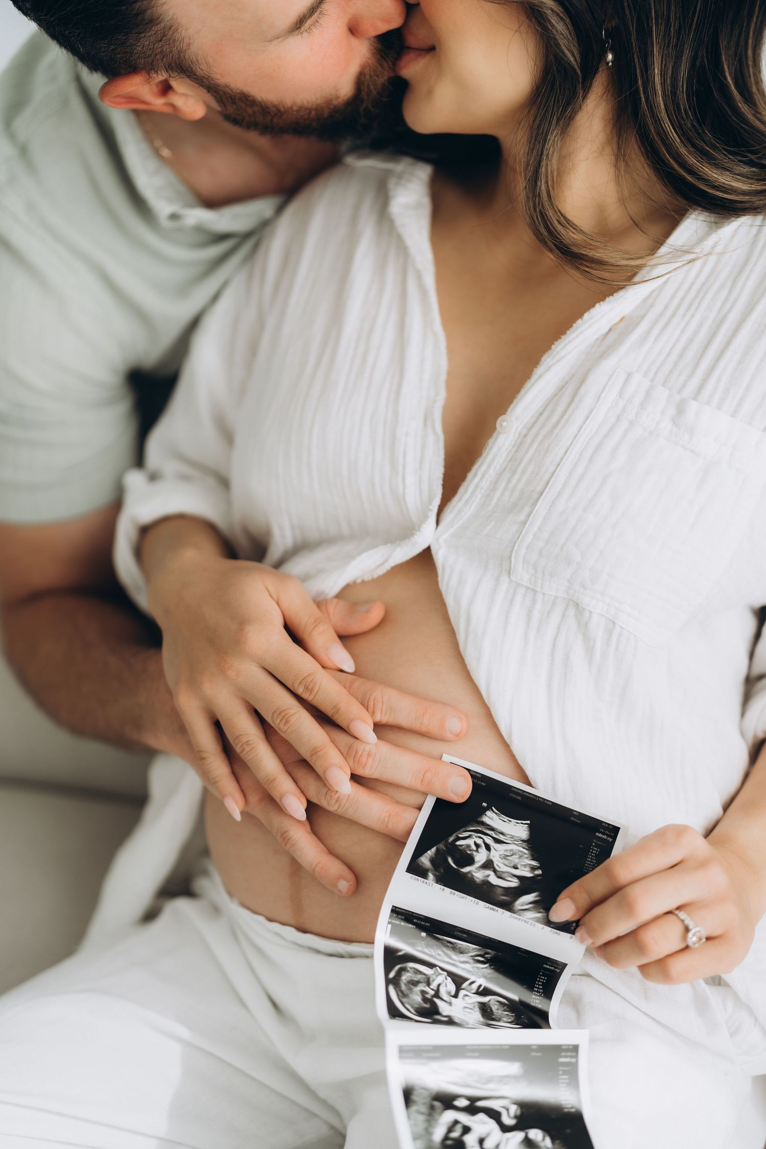 Couple in white clothing holding ultrasound photos while lying on bed in intimate maternity photoshoot.