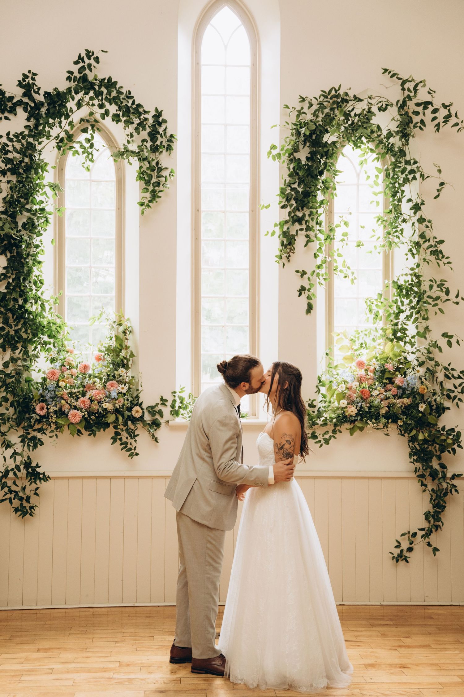 Romantic wedding couple embraces in a light-filled room with climbing vines and windows creating a dreamy atmosphere.