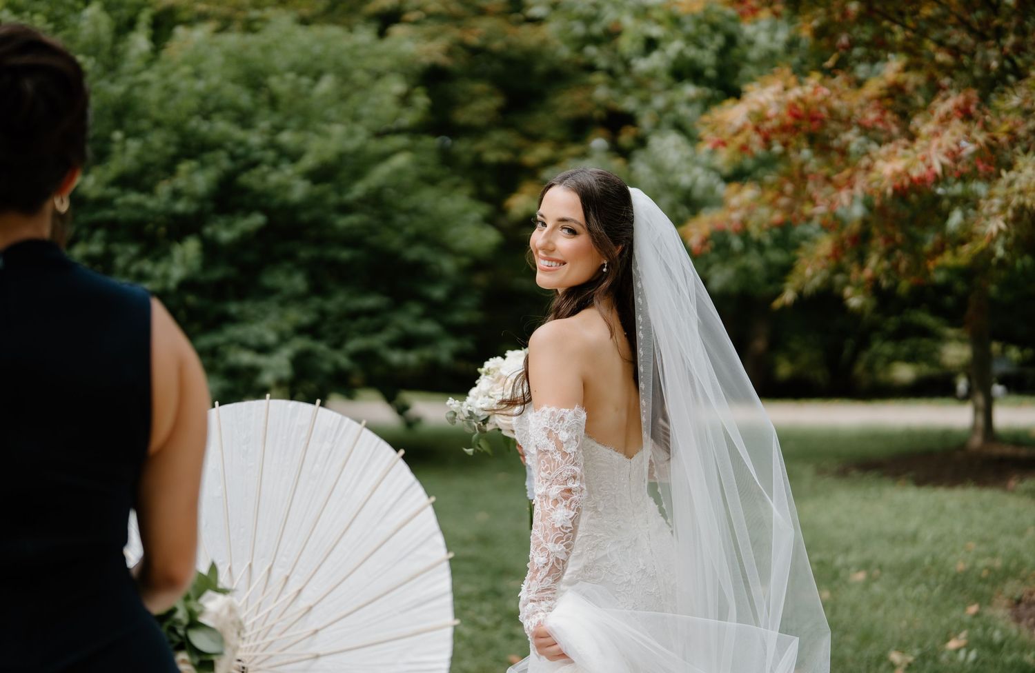 A bride in a white wedding dress poses with a delicate white parasol in a lush garden setting.