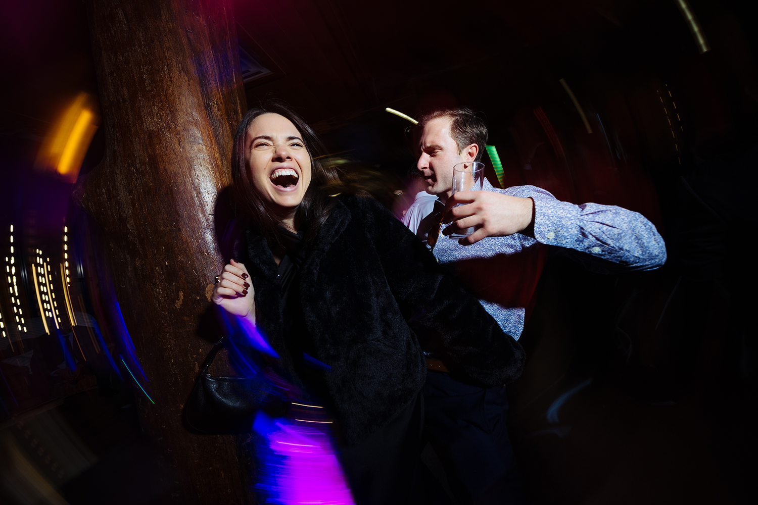 Two people dancing energetically at a wedding reception with colorful lights in the dark background and shutter drag effect.