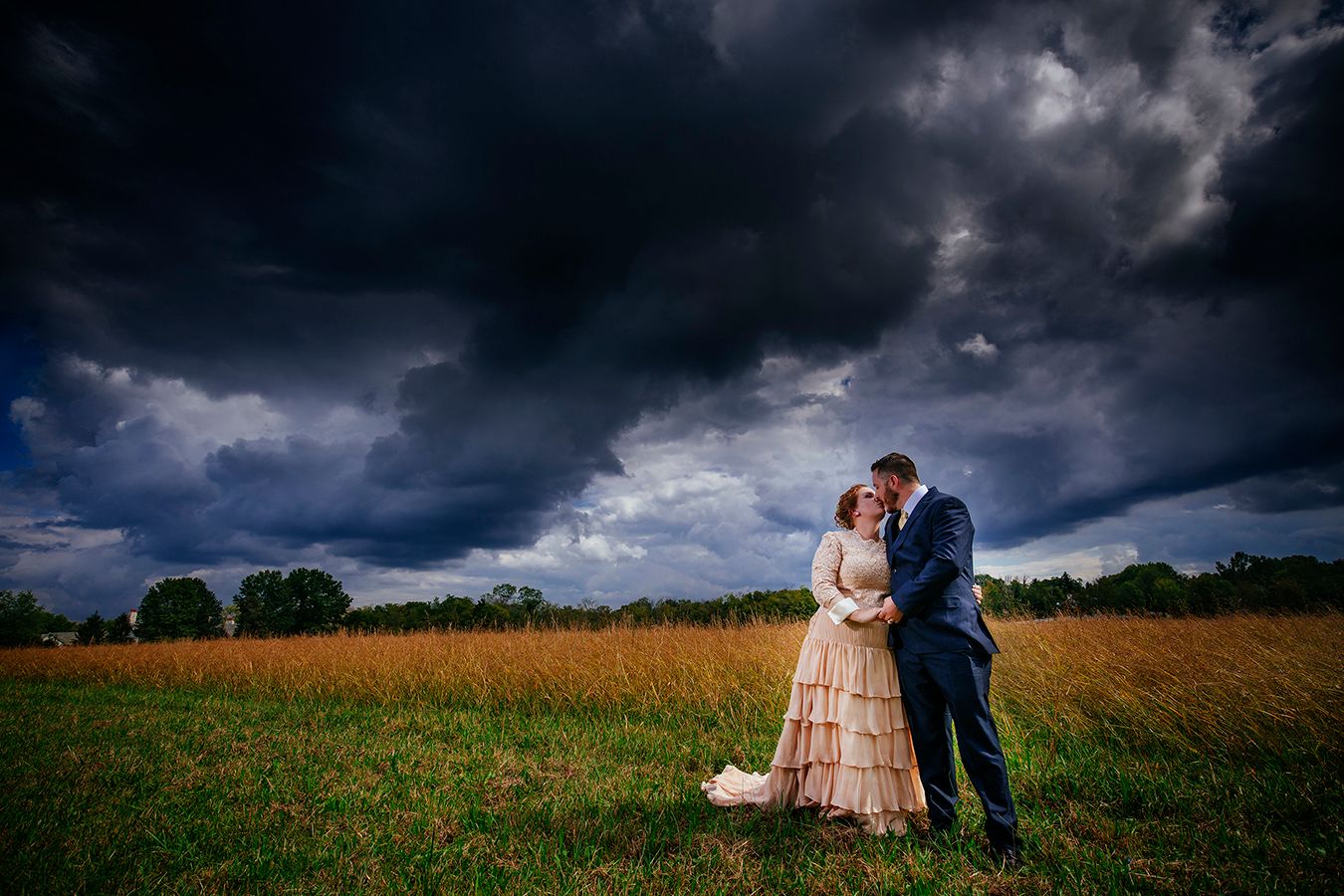 A couple embraces in a golden field under dramatic storm clouds during a romantic outdoor photo session.