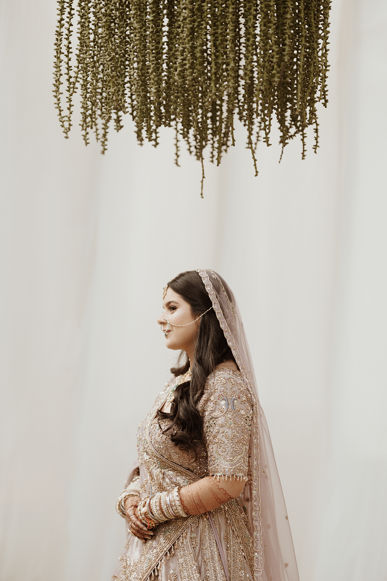 Side profile of an indian bride in an ornate beige gown and veil standing beneath hanging floral decor.
