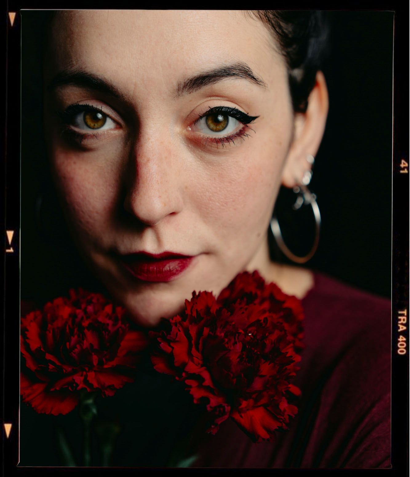 Woman with bold eyeliner and red lips holds red carnations close to her face in a dark, moody film-style portrait.
