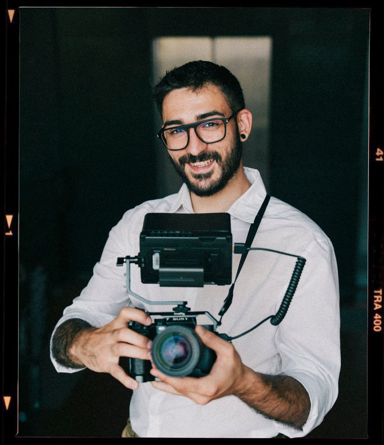 Smiling man with glasses holding a professional film camera with flash attachment, wearing a white shirt.