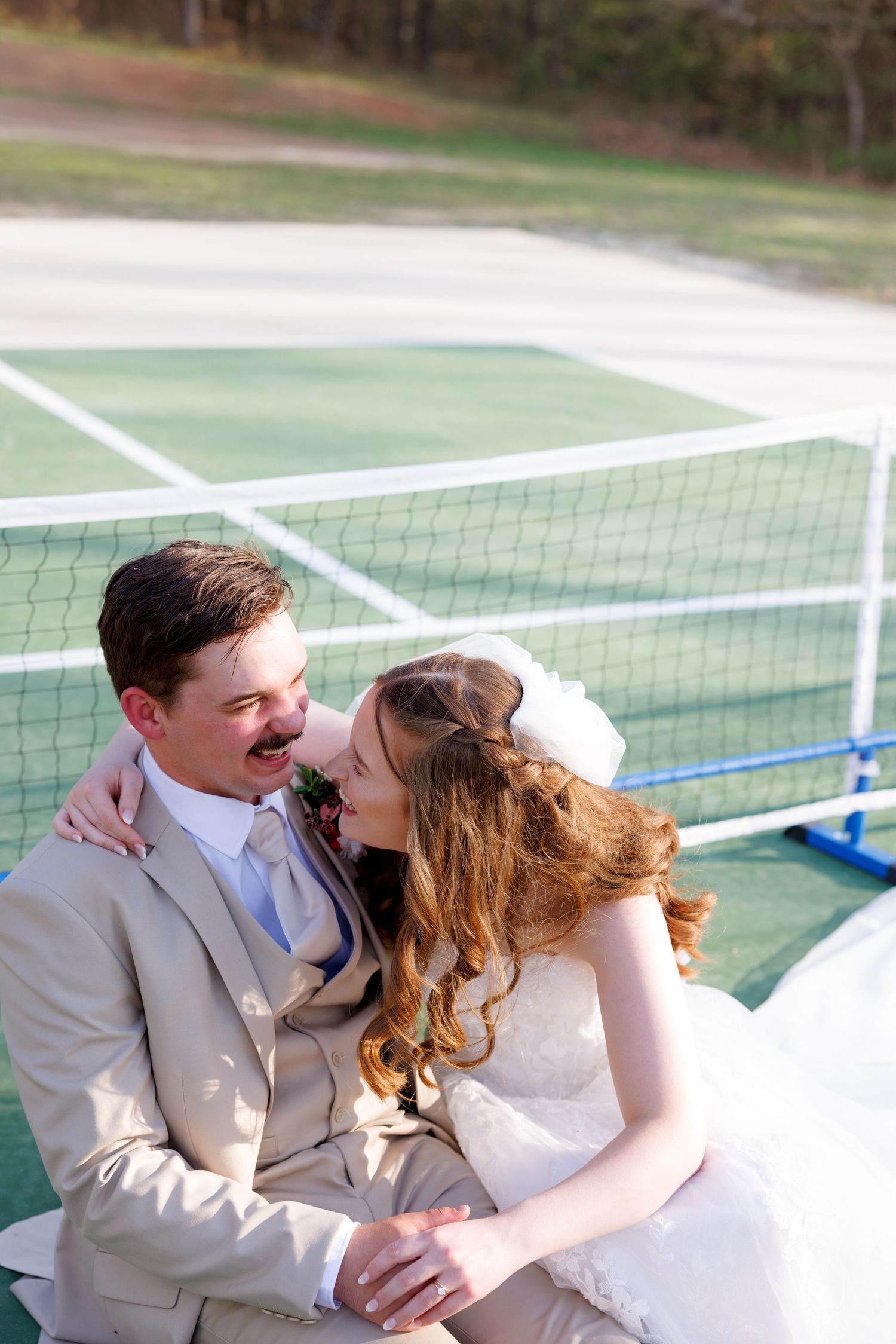 Bride and groom share a romantic moment on a tennis court, with the bride in white and groom in a tan suit.
