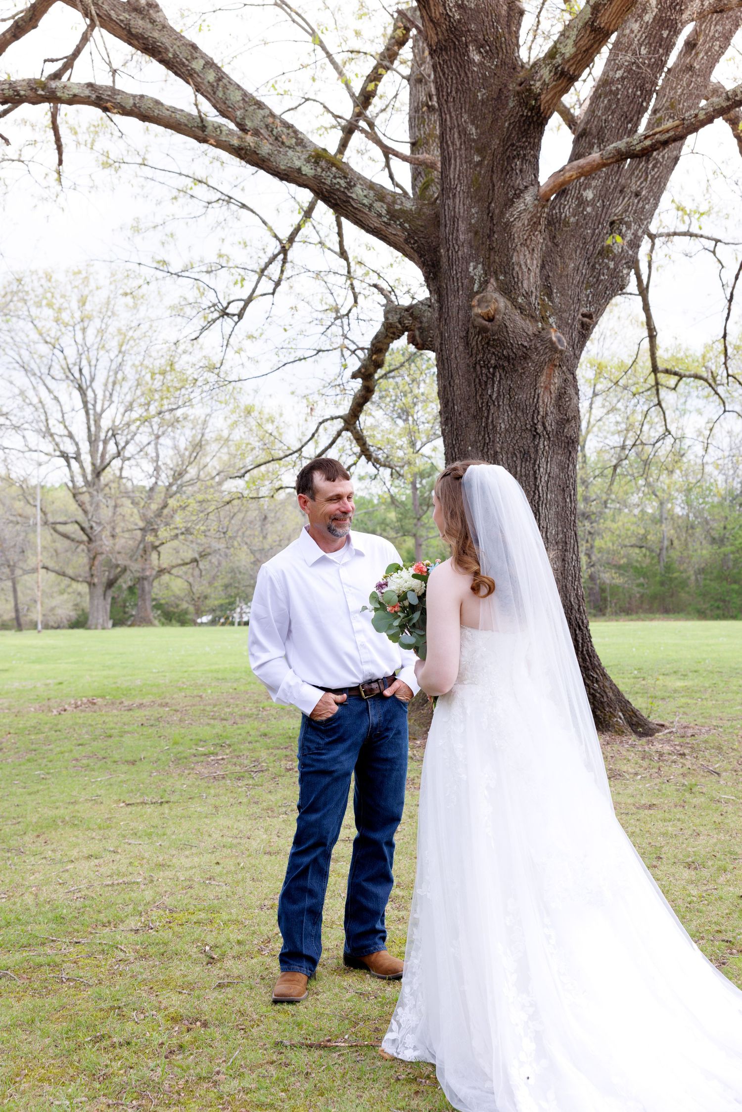 Bride in white lace gown and veil holds bouquet facing groom in jeans under large oak tree at outdoor wedding.