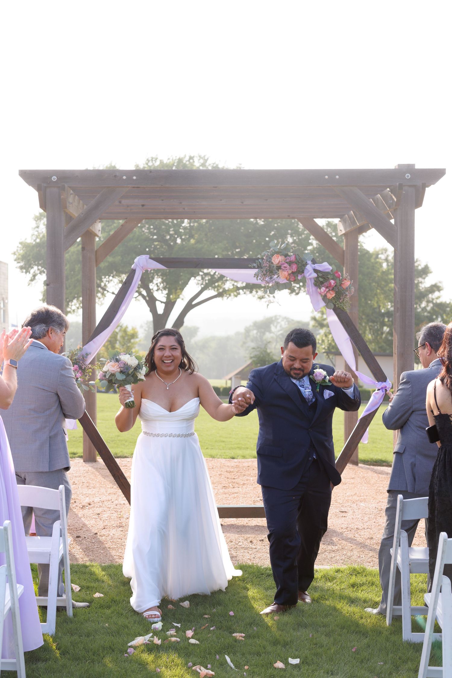 Happy newlyweds walk back down the aisle under a rustic wooden arch as guests celebrate their outdoor wedding.