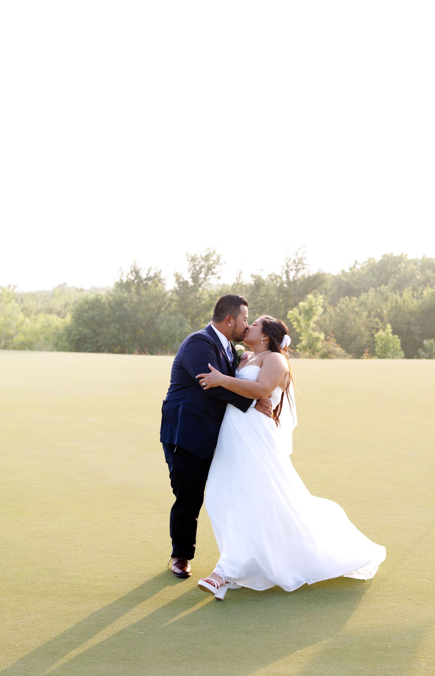 Bride and groom share a romantic kiss on a lush green golf course in golden hour light, bride in white gown.