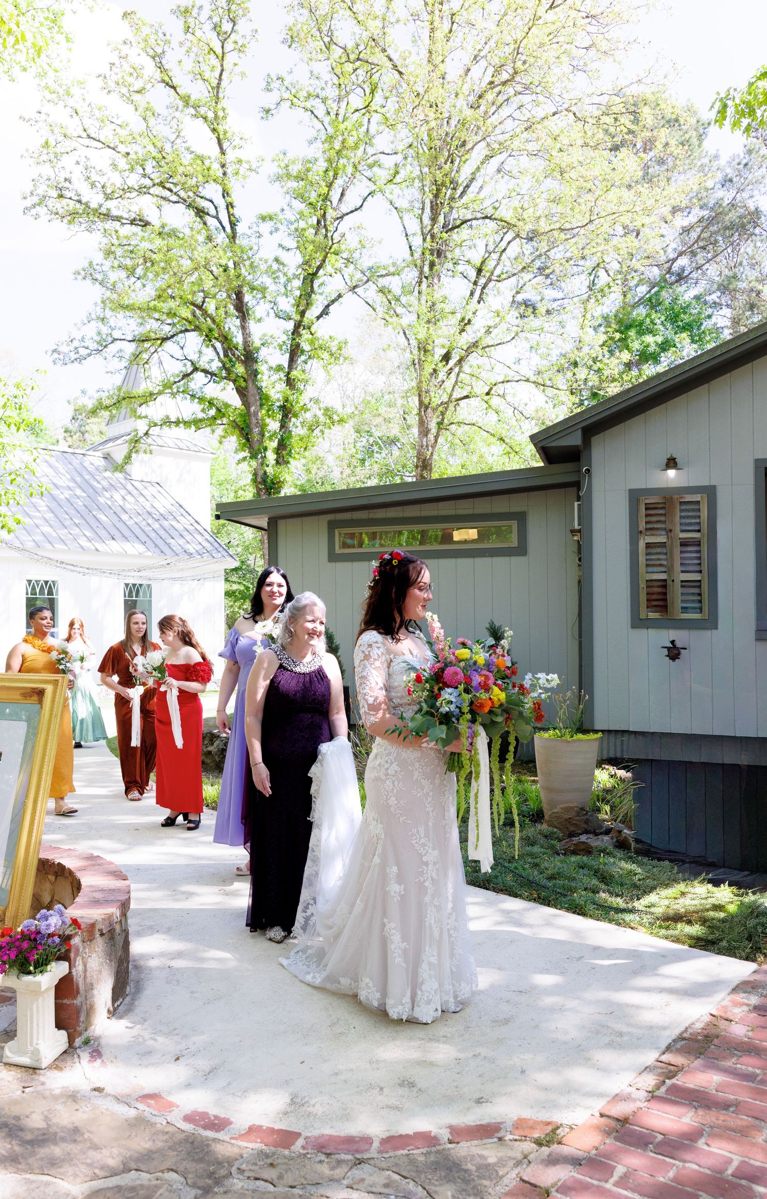 Bride in white lace gown walks with bridesmaids in colorful dresses during outdoor wedding ceremony.