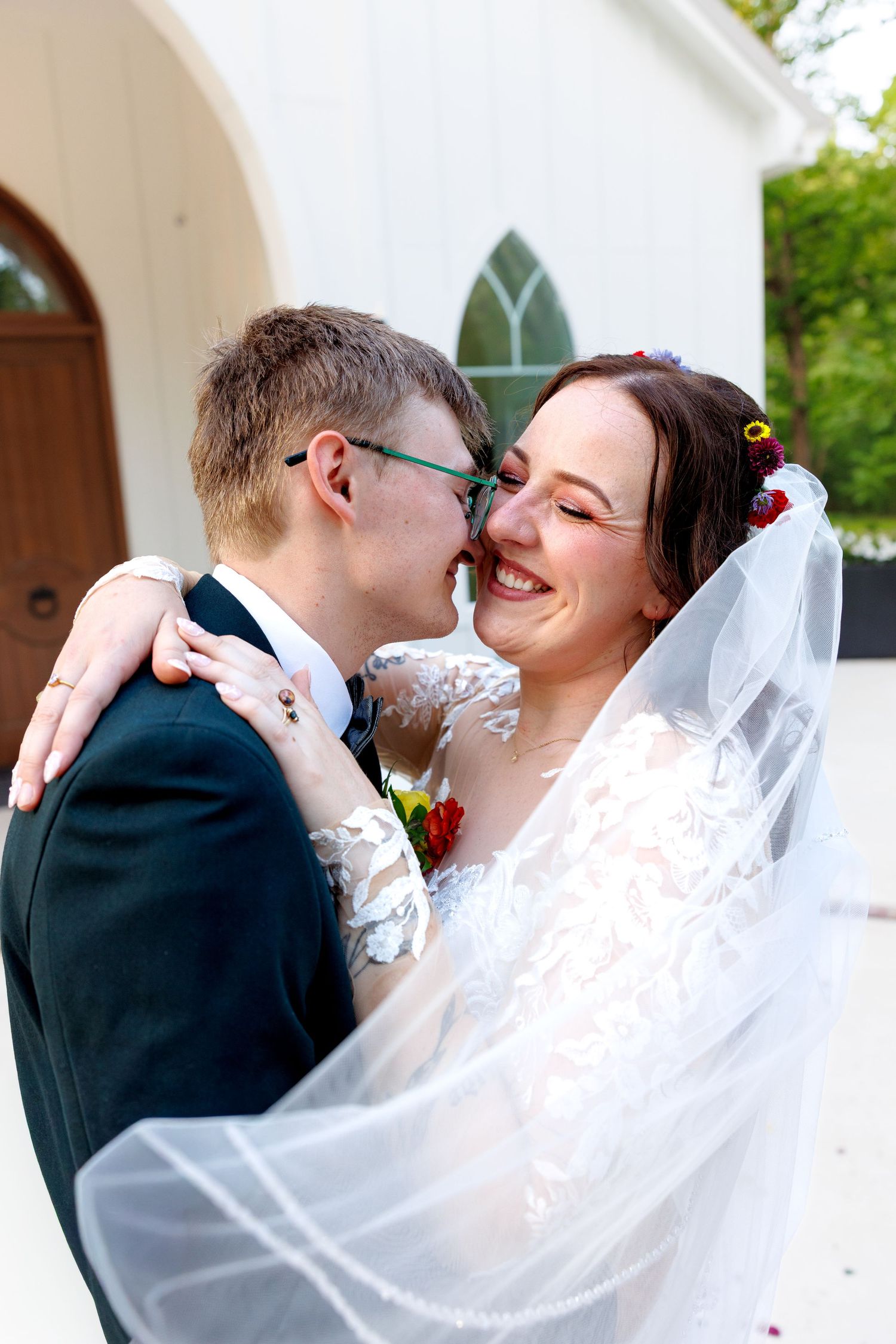 Smiling bride in lace gown and veil embraces groom in navy suit with green glasses outside a white chapel.
