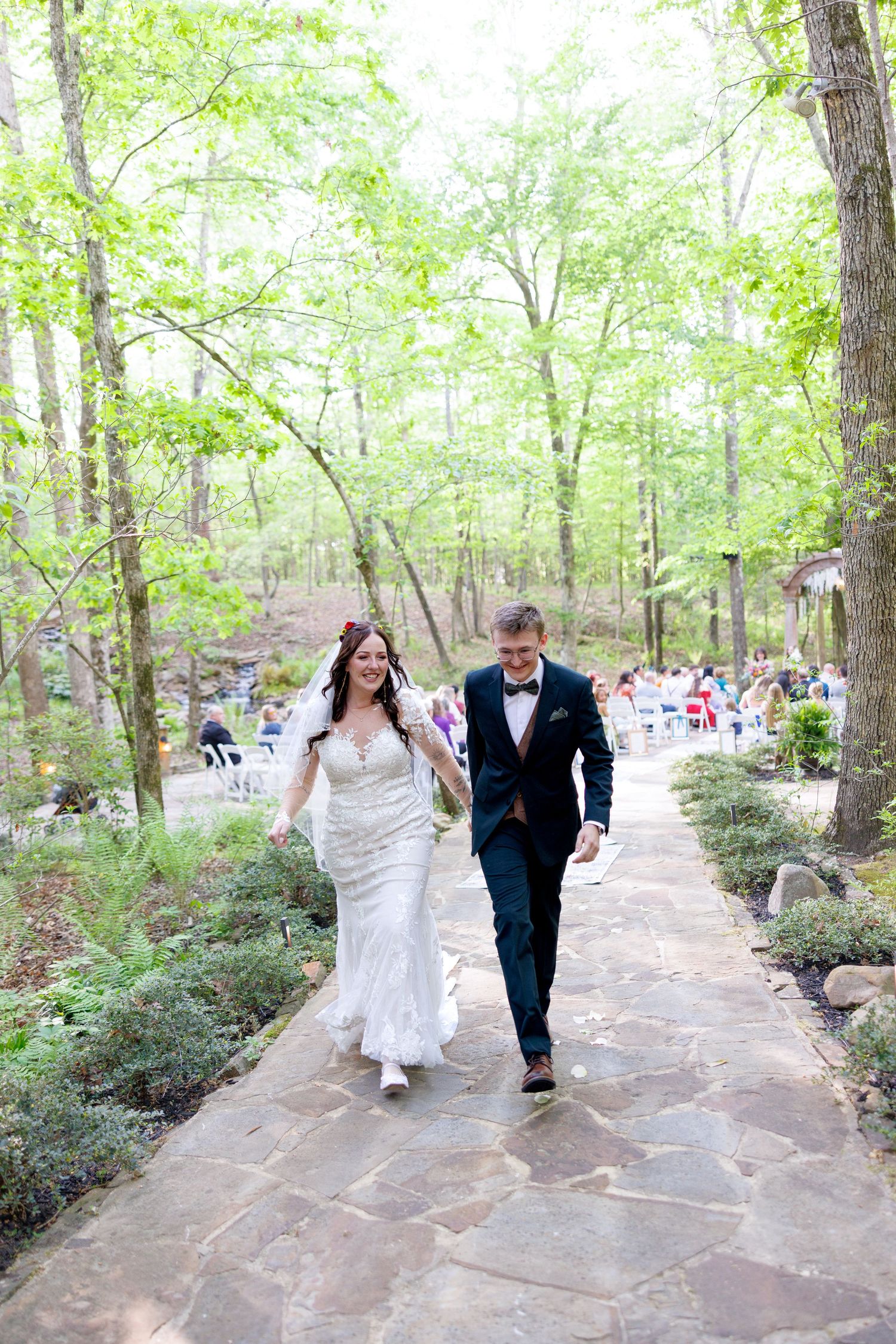 A bride and groom walk hand-in-hand down a stone path through lush green woods after their ceremony.