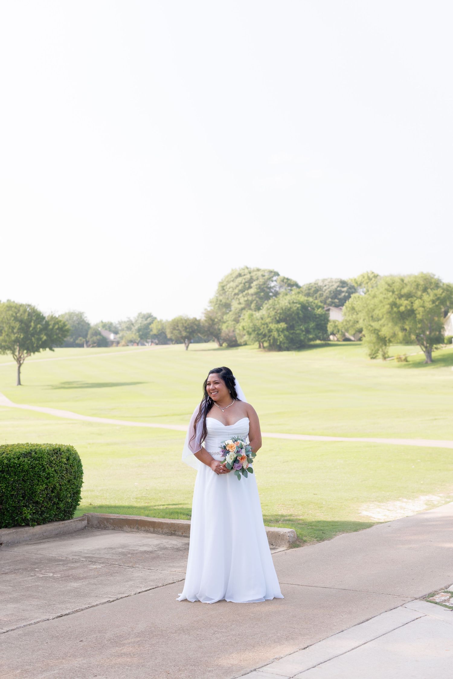 Bride in strapless white gown holds colorful bouquet while posing on golf course pathway on wedding day.