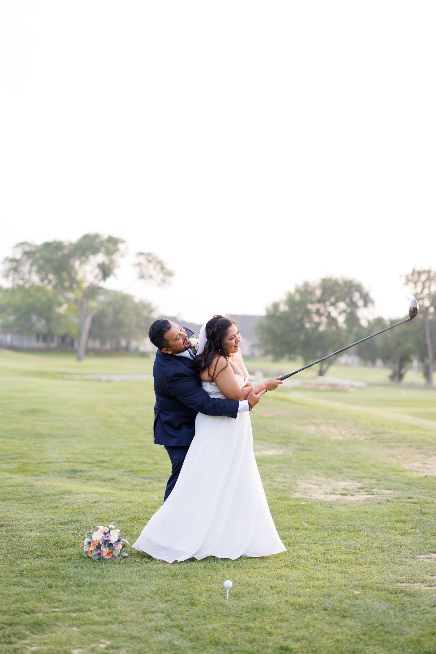 Groom helps bride swing a golf club on a lush golf course during their wedding photo session.