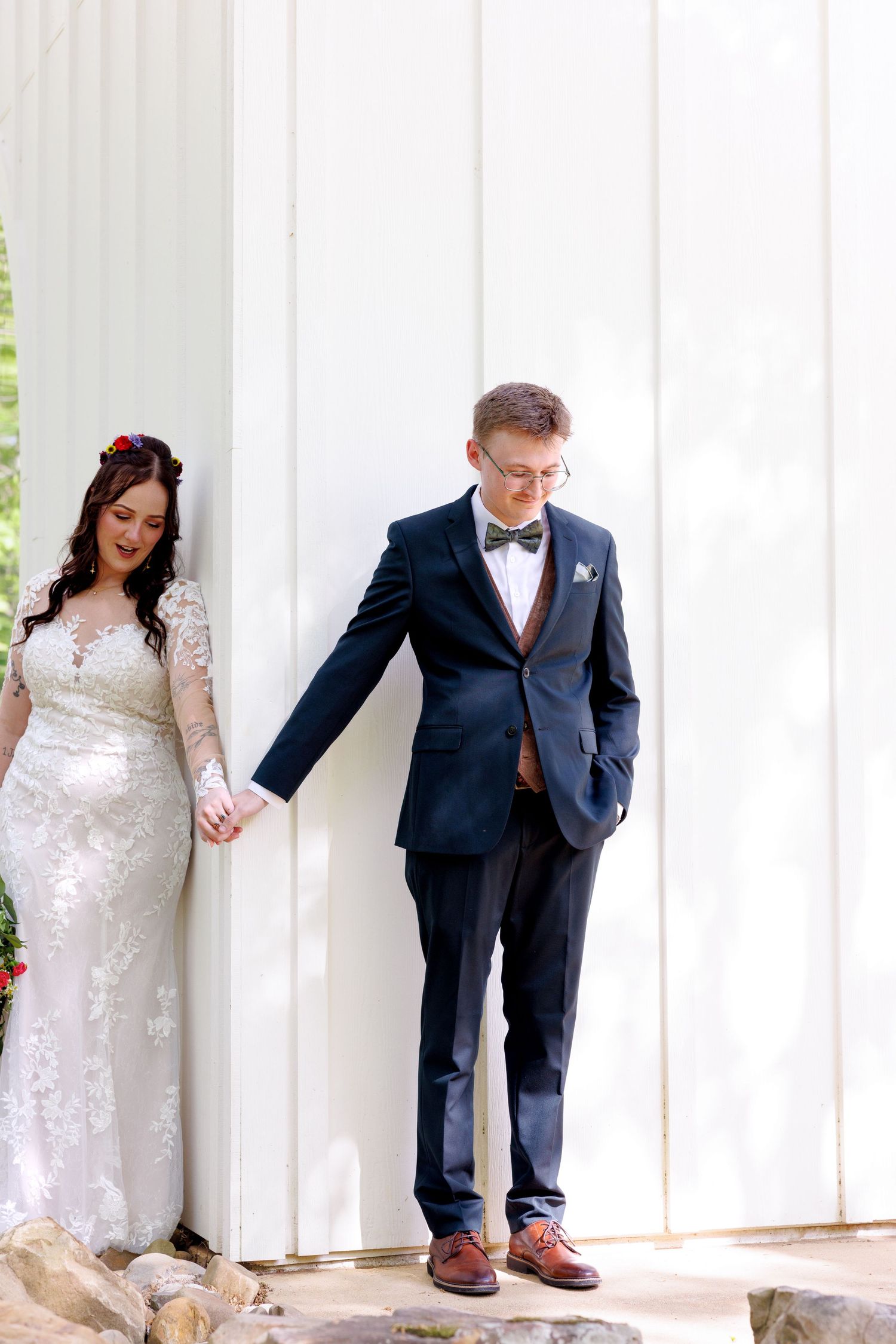 Bride in lace gown and groom in navy suit holding hands against a white barn wall on their wedding day.