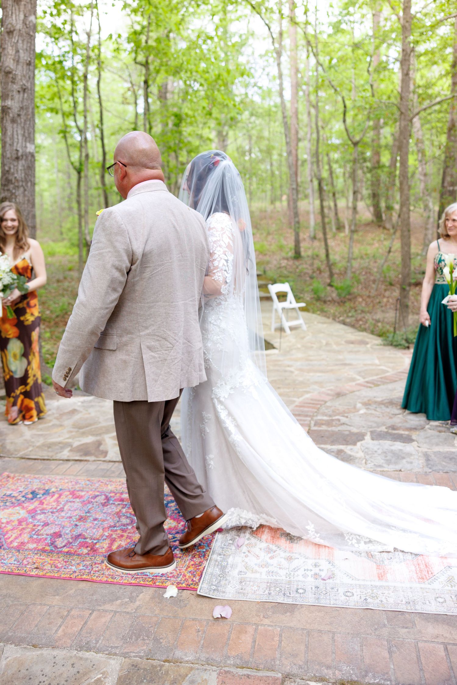 Bride in lace gown with long veil walks with escort on colorful rug at outdoor woodland wedding ceremony.