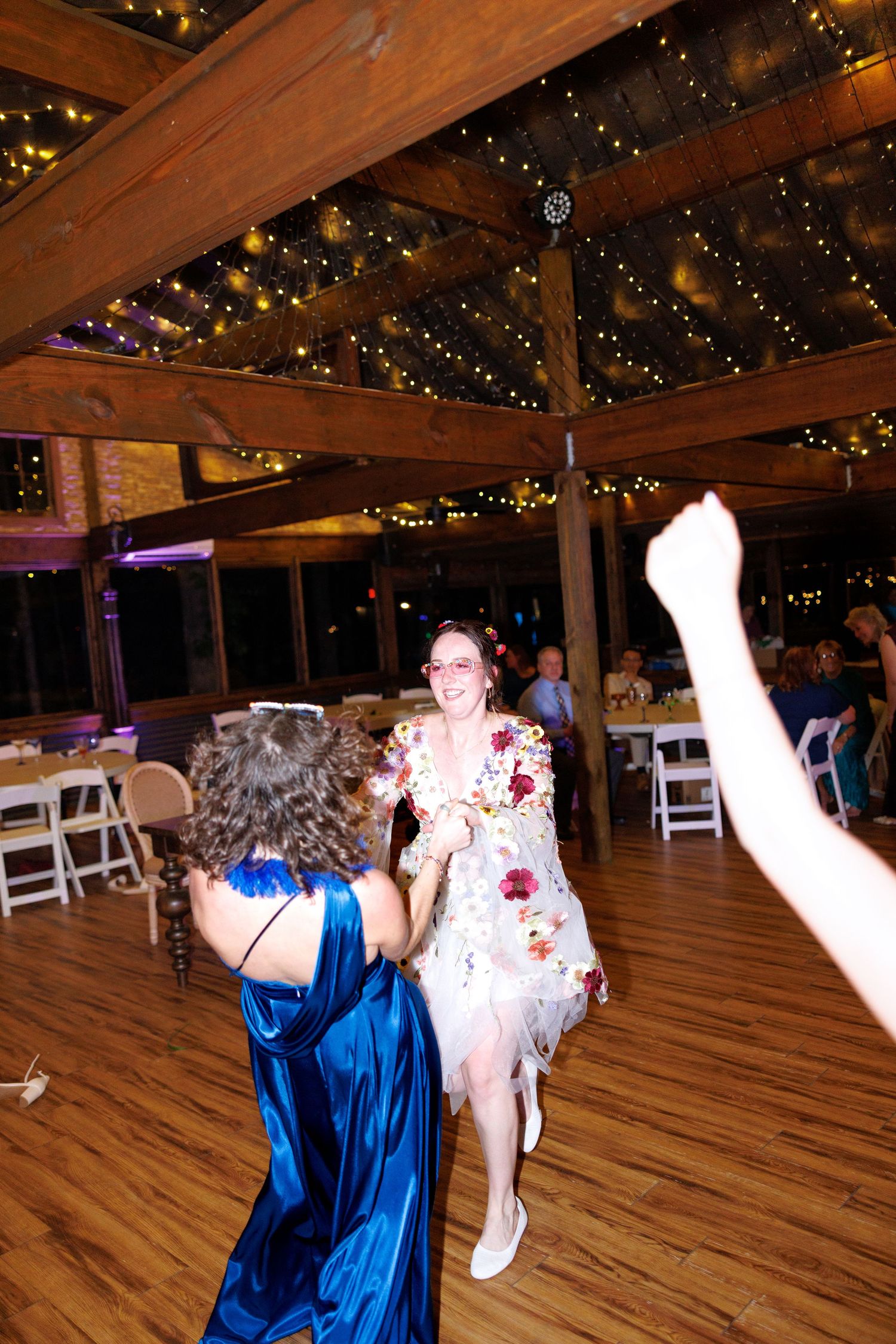 Two women dancing joyfully at a rustic wedding reception with string lights and wooden beams overhead.