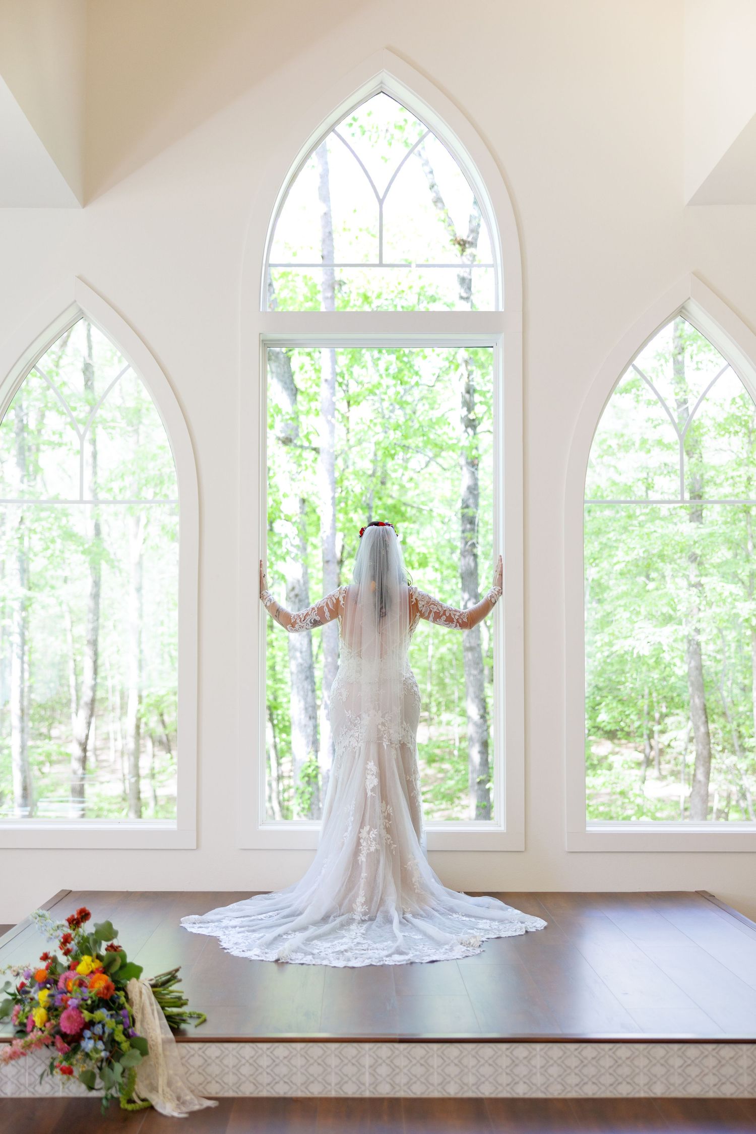 Bride in lace wedding gown and veil stands before gothic arched windows in a white chapel overlooking lush green trees.
