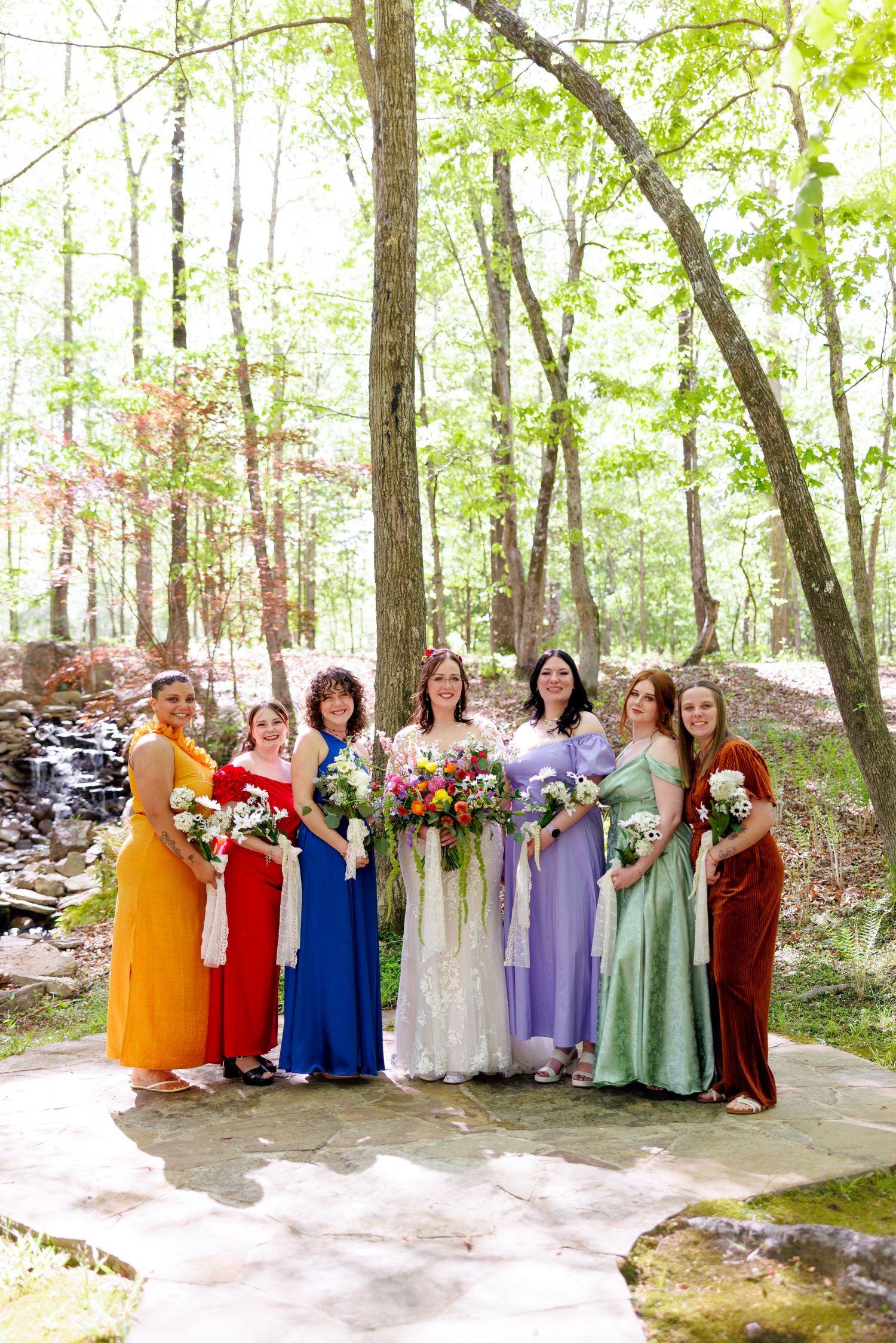 Bride and six bridesmaids in colorful rainbow dresses pose together in a lush forest wedding setting.