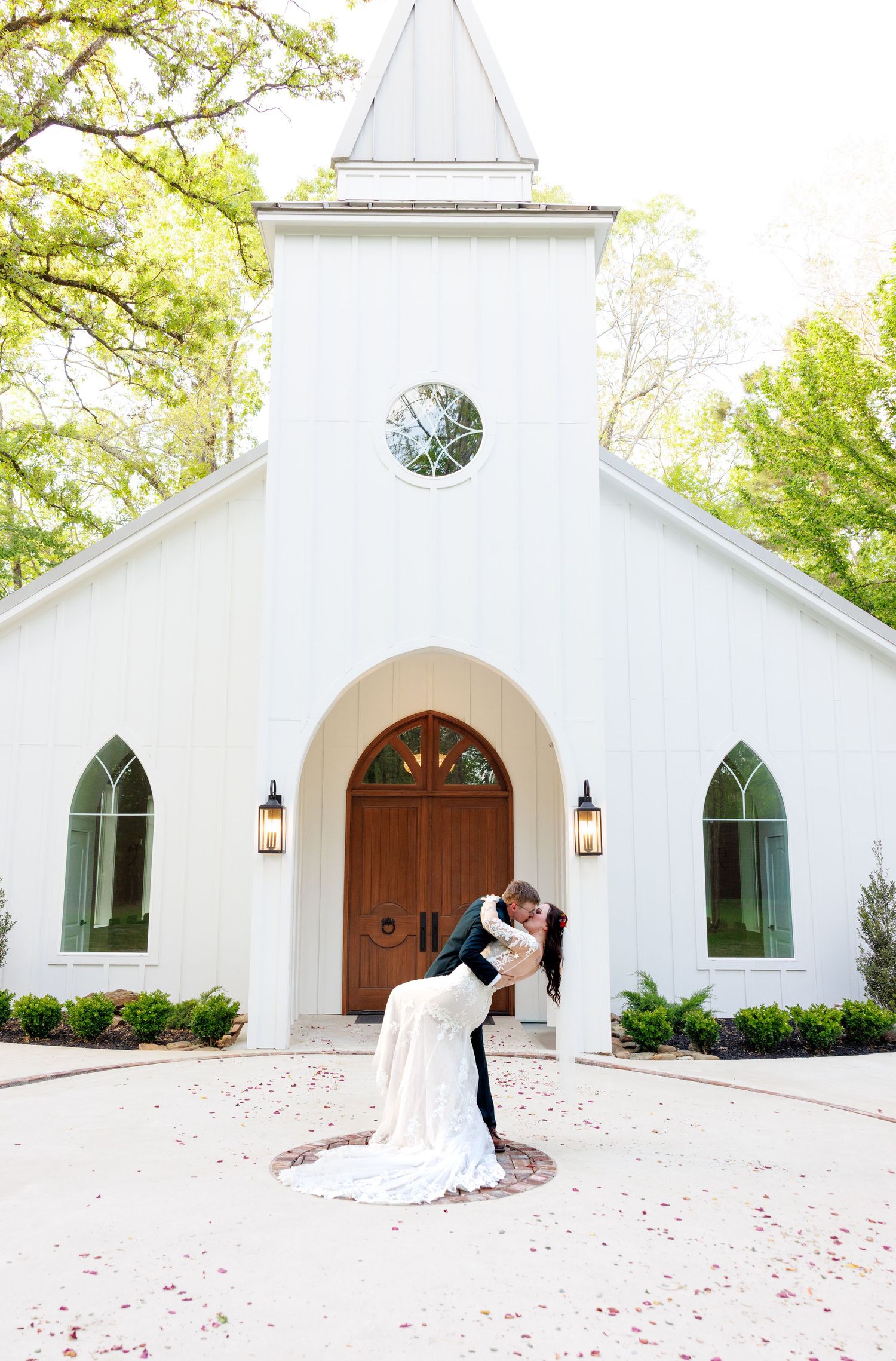 Groom dips bride in a romantic pose in front of a charming white wedding chapel with a steeple and arched doors.