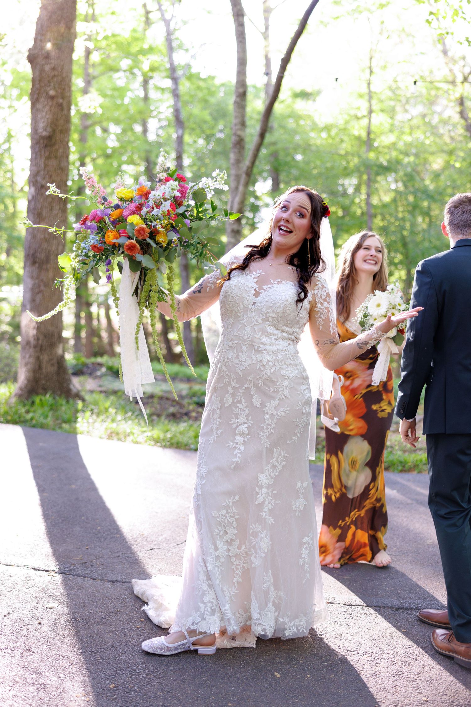 Joyful bride in lace wedding gown tosses colorful bouquet outdoors surrounded by wedding guests.