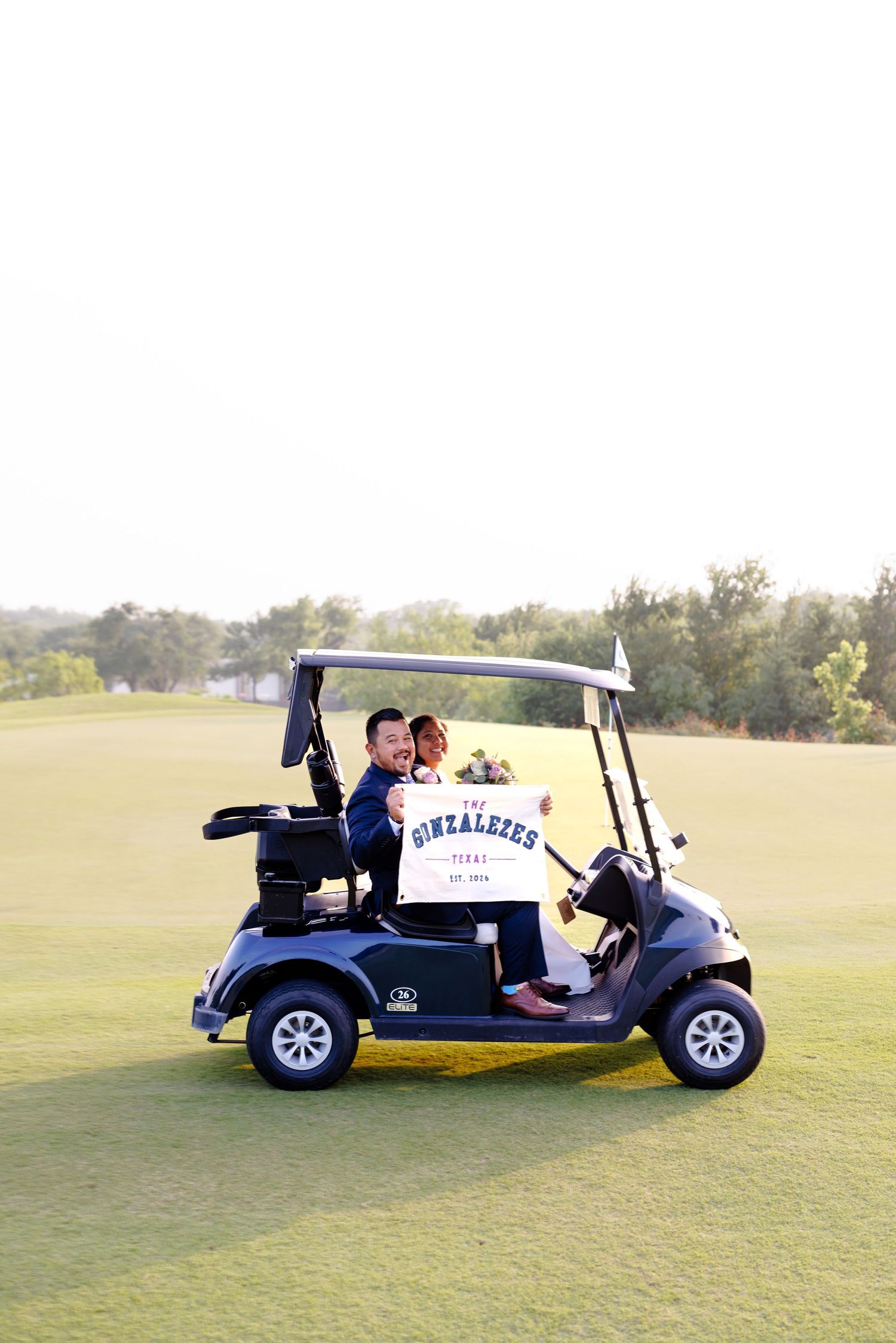 Newlyweds celebrate on a golf cart holding a Gonzalezes sign on a sunny green golf course.
