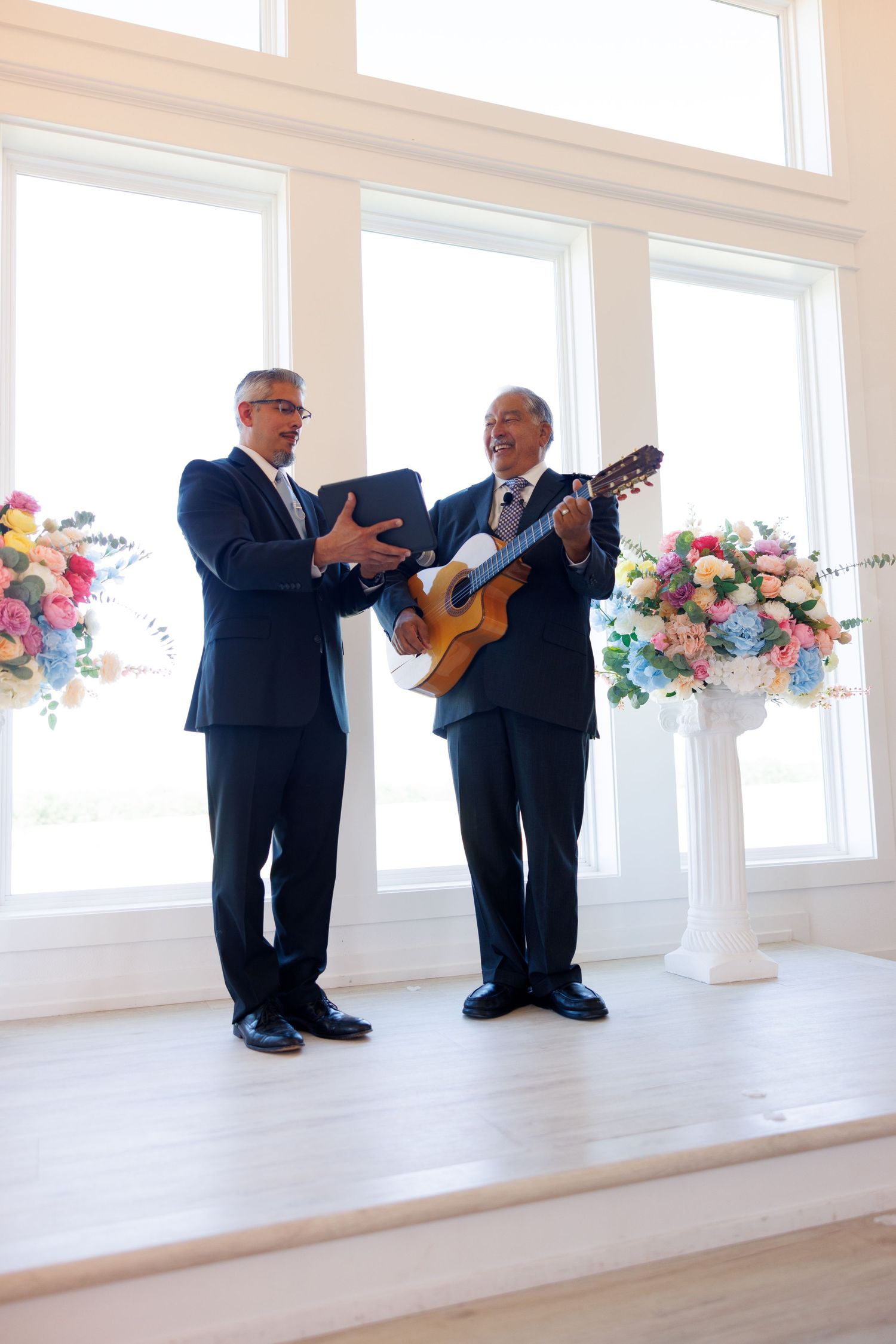 Two musicians in suits perform with guitars in bright venue with floral decorations.