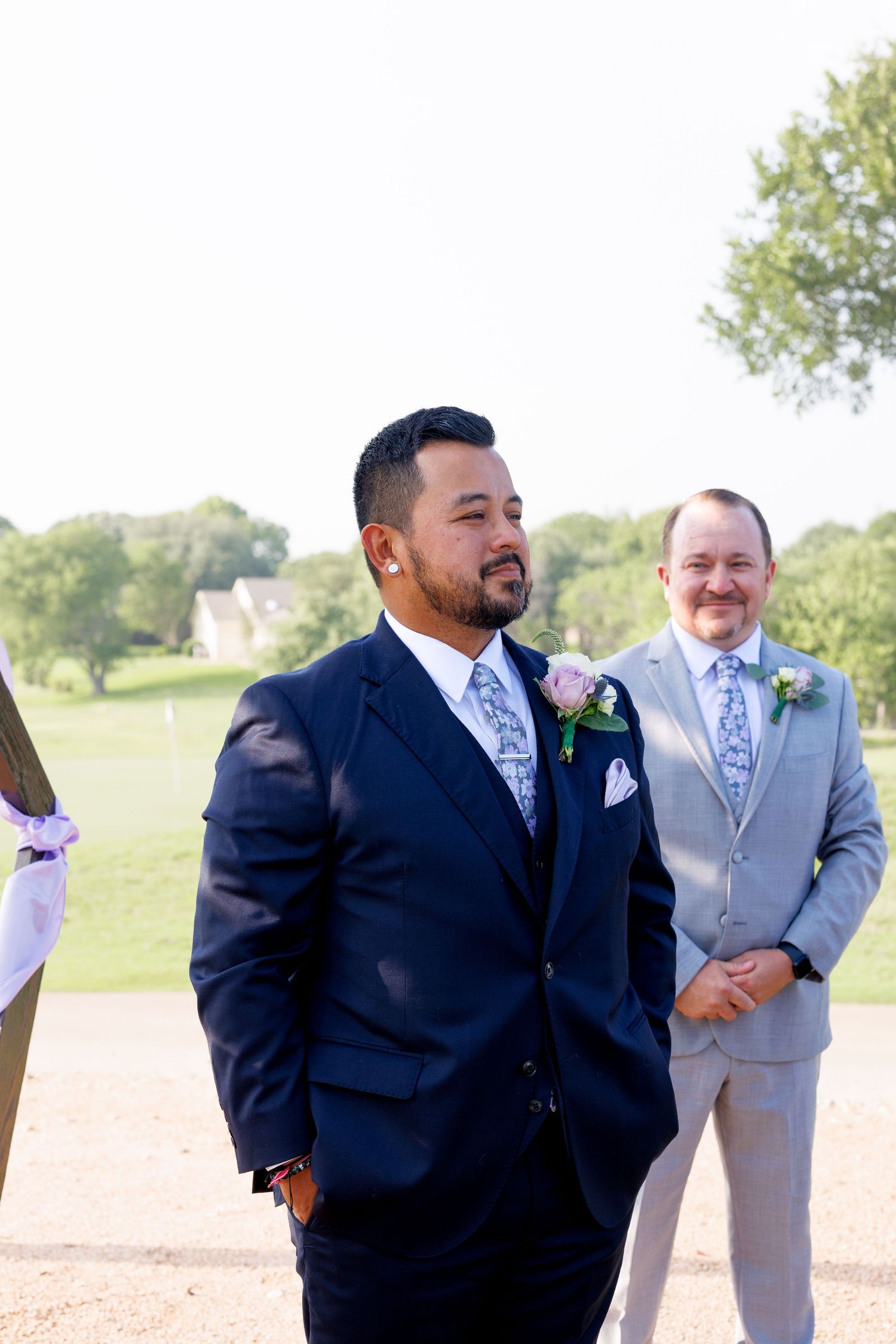 Two groomsmen in navy and gray suits with floral boutonnieres stand outdoors at a wedding ceremony.