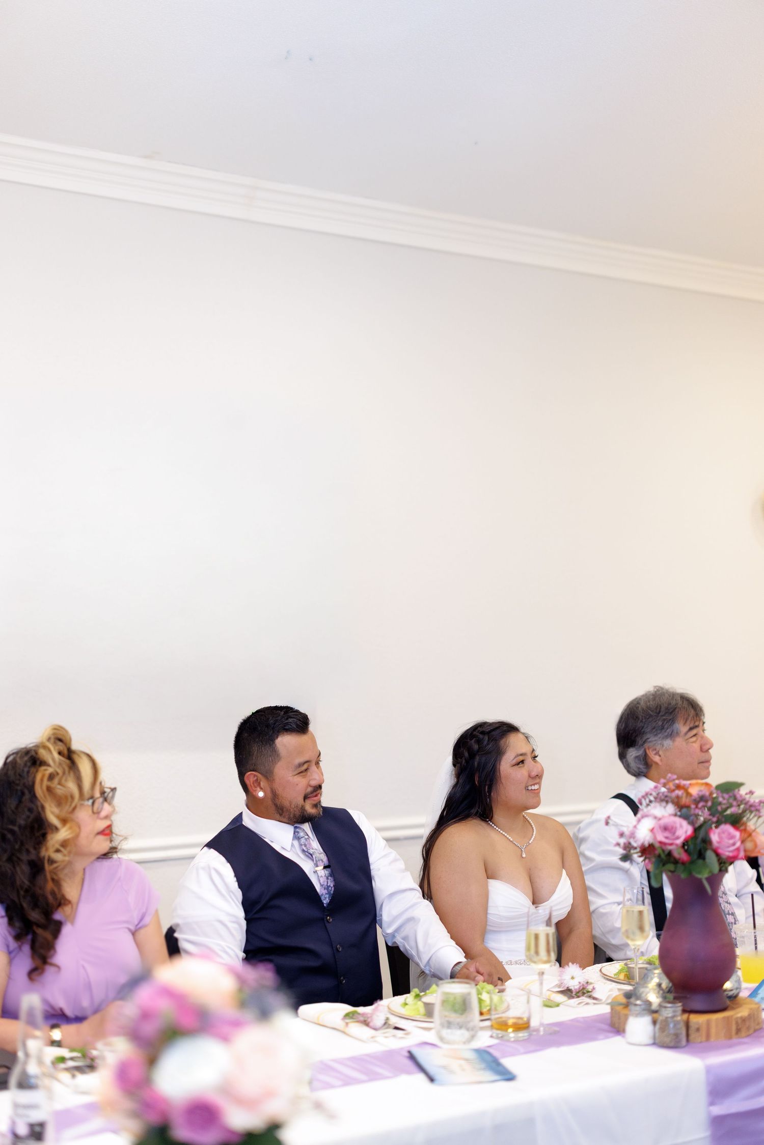Bride and groom laughing at wedding reception head table with family, purple floral centerpieces and lavender decor.