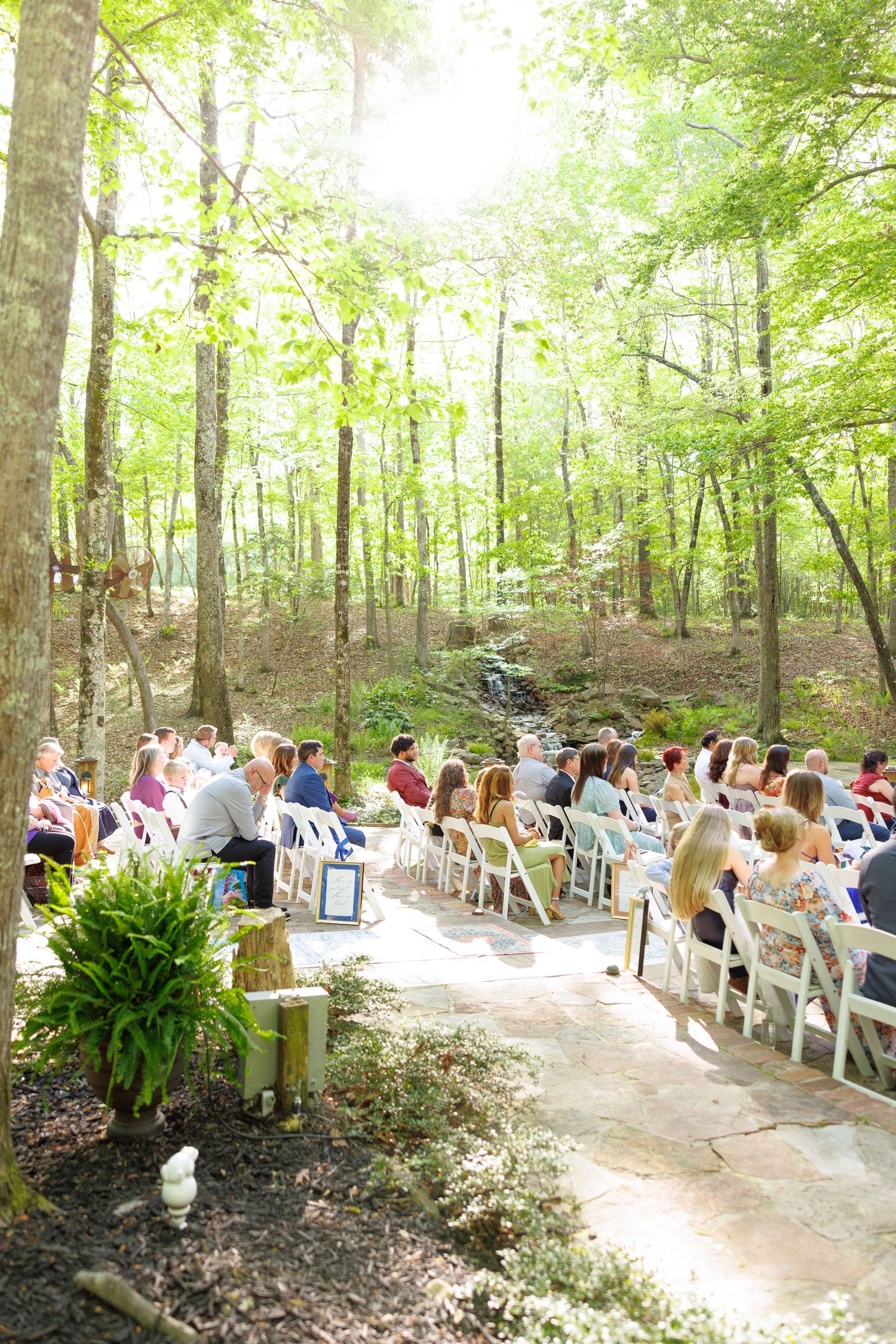 Outdoor woodland wedding ceremony with guests seated in white chairs among tall trees and dappled sunlight.