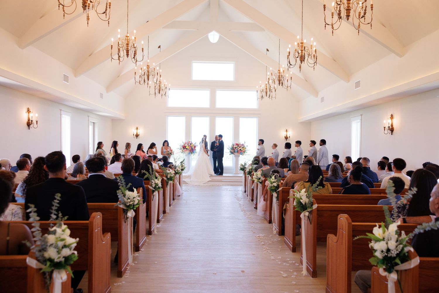 Wide view of wedding chapel with wooden pews, chandeliers, and natural light streaming through windows.