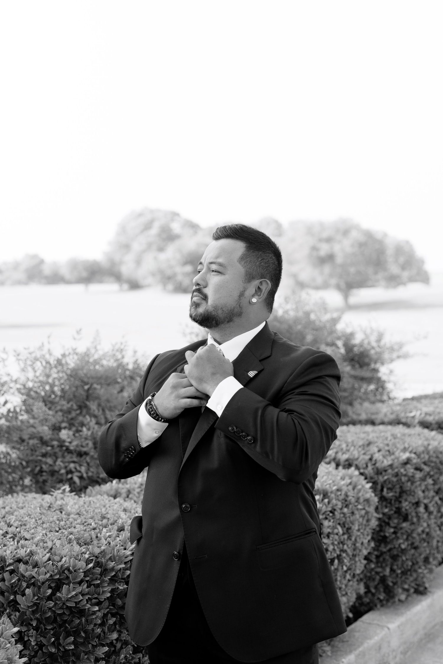 Black and white portrait of a well-dressed man in a suit adjusting his tie outdoors near manicured hedges.