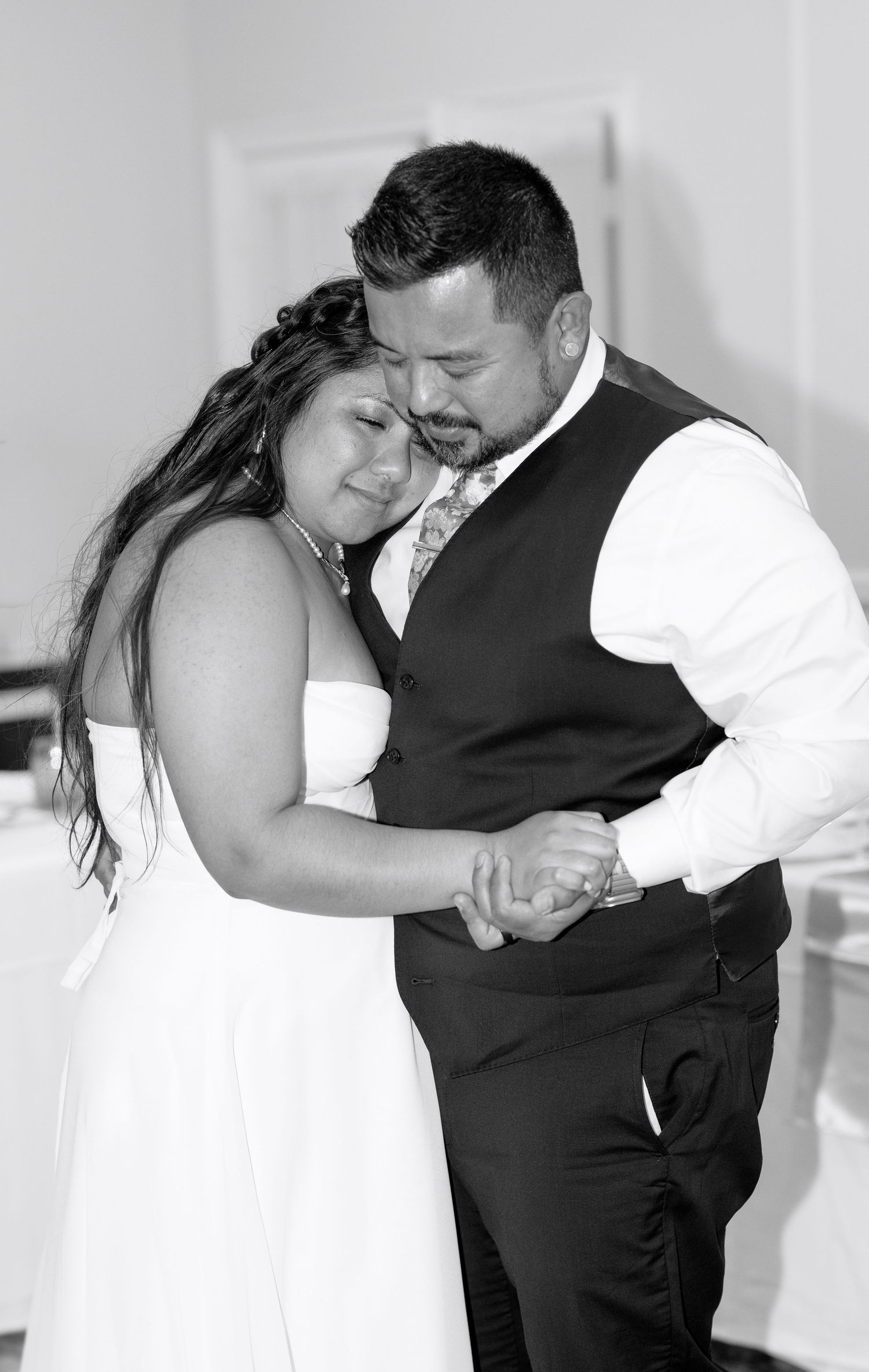 Black and white photo of a bride and groom sharing their first dance, holding hands and embracing at their wedding reception.