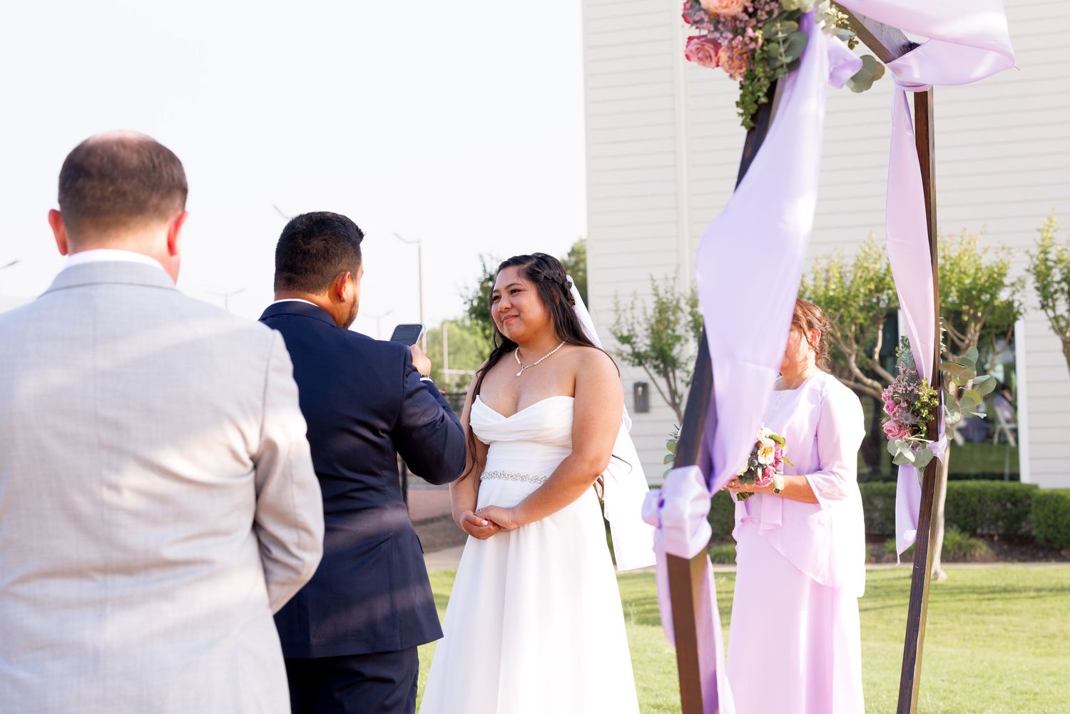 Outdoor wedding ceremony with bride in white gown and groom in navy suit exchanging vows under floral arch.