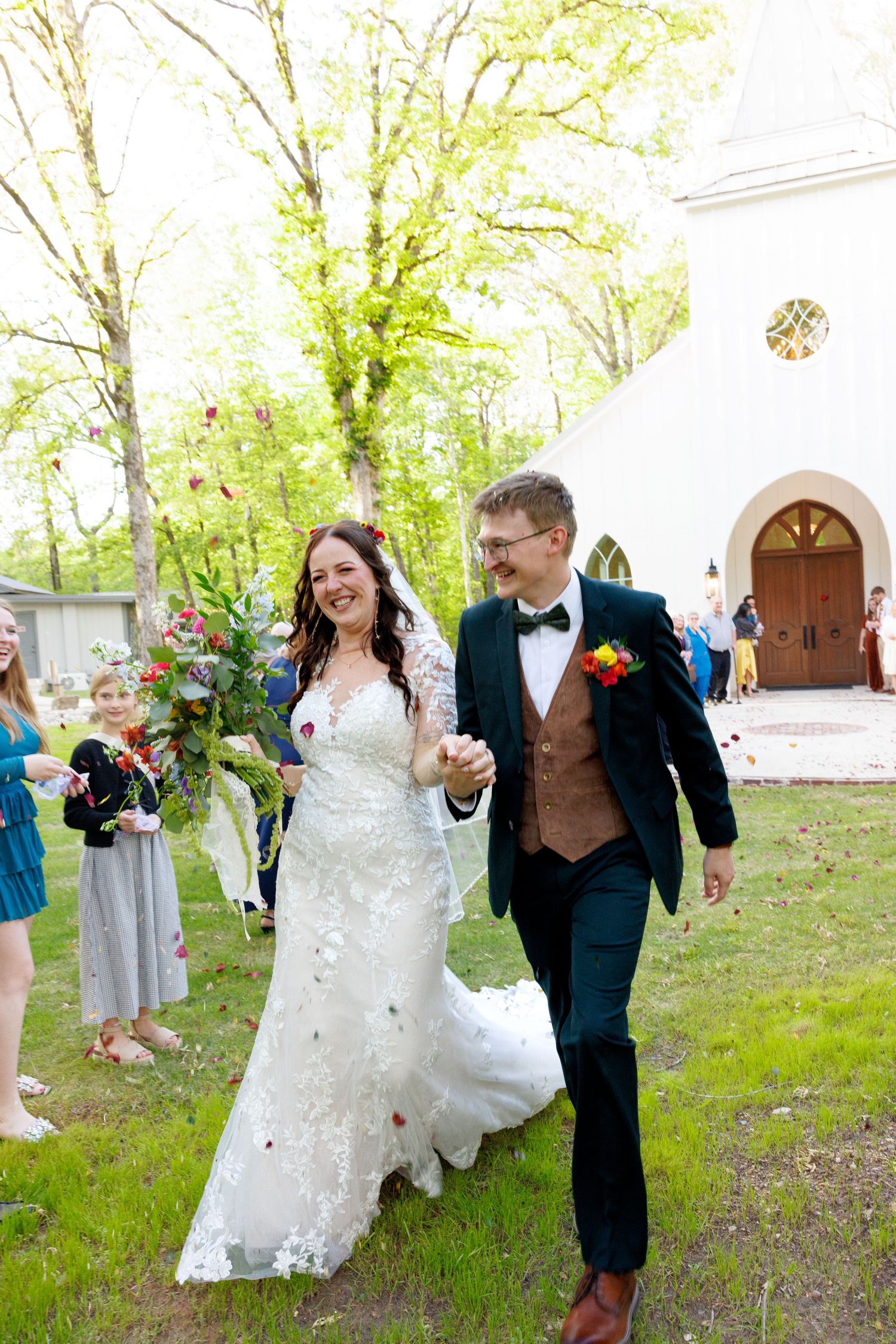 Happy newlyweds walking hand-in-hand outside white church after wedding ceremony with guests celebrating.
