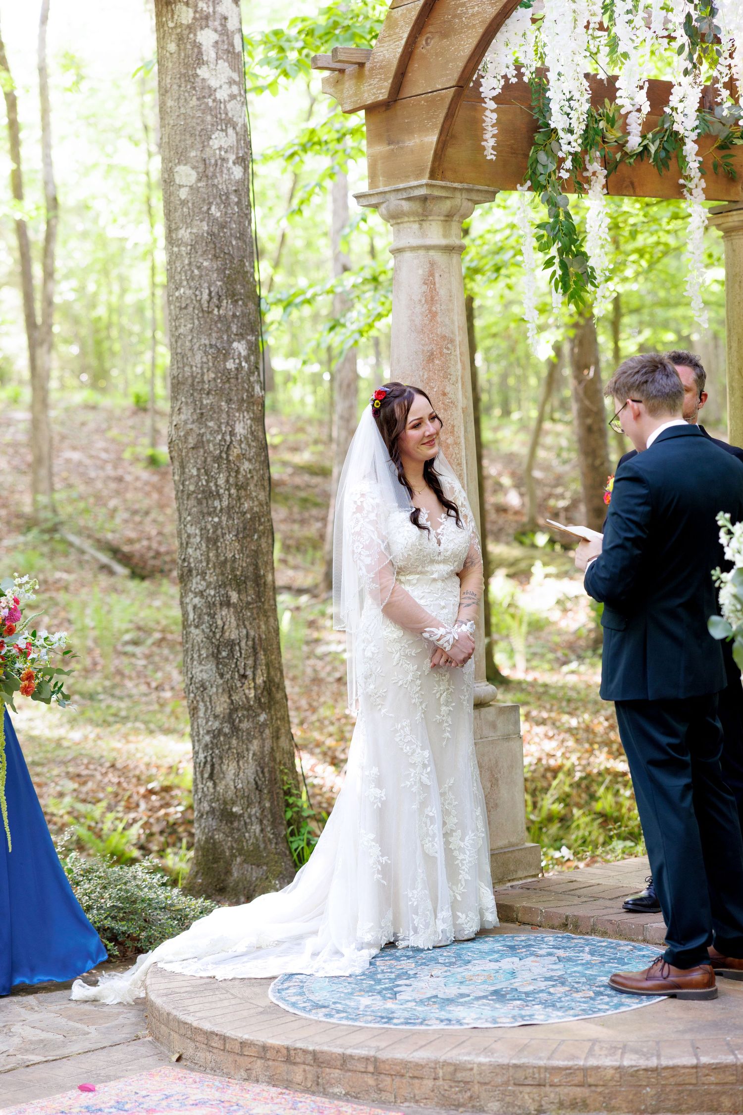 Bride in lace gown and veil smiles during outdoor woodland wedding ceremony under stone gazebo.