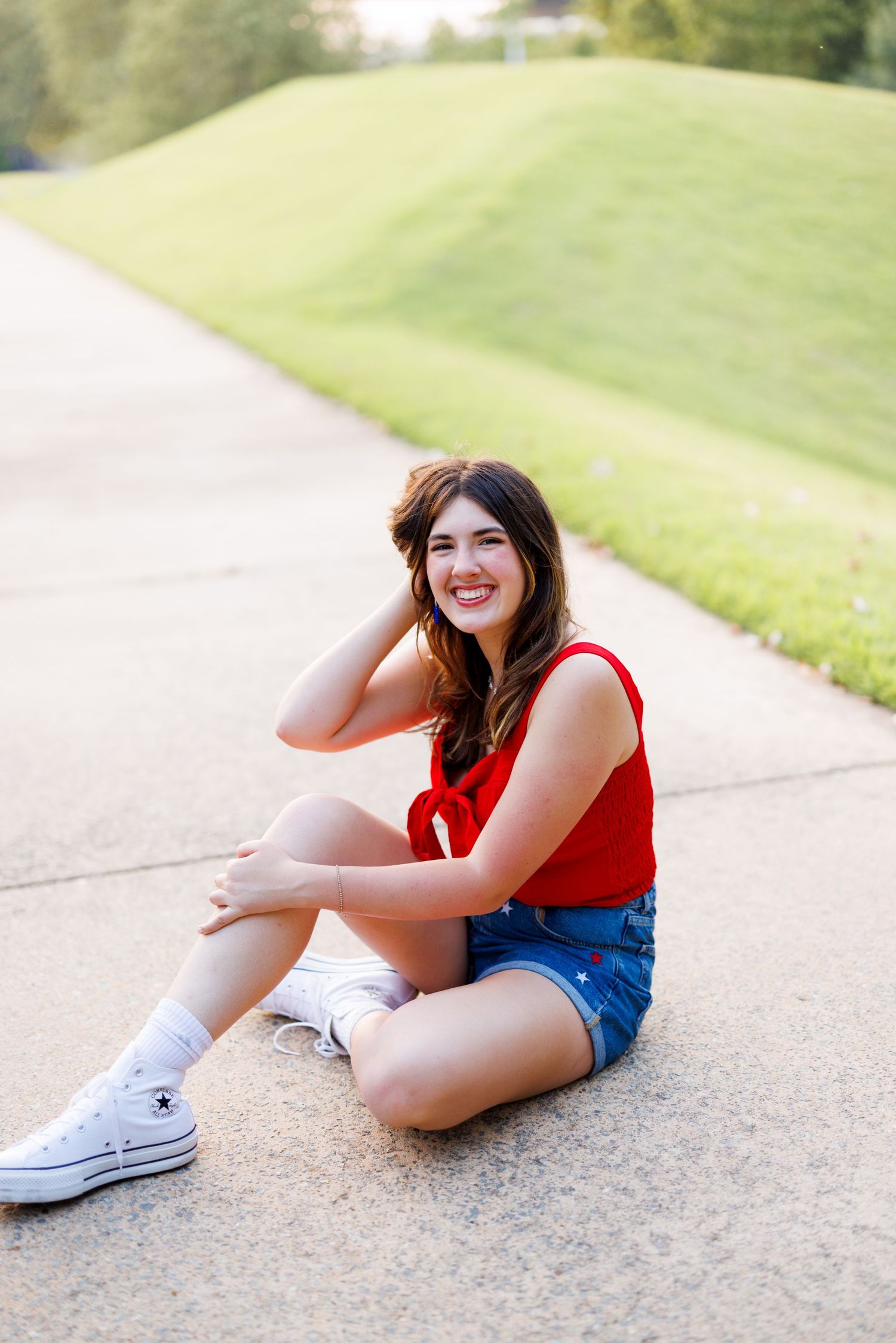 Person in red top and shorts sits on concrete path during outdoor photoshoot.