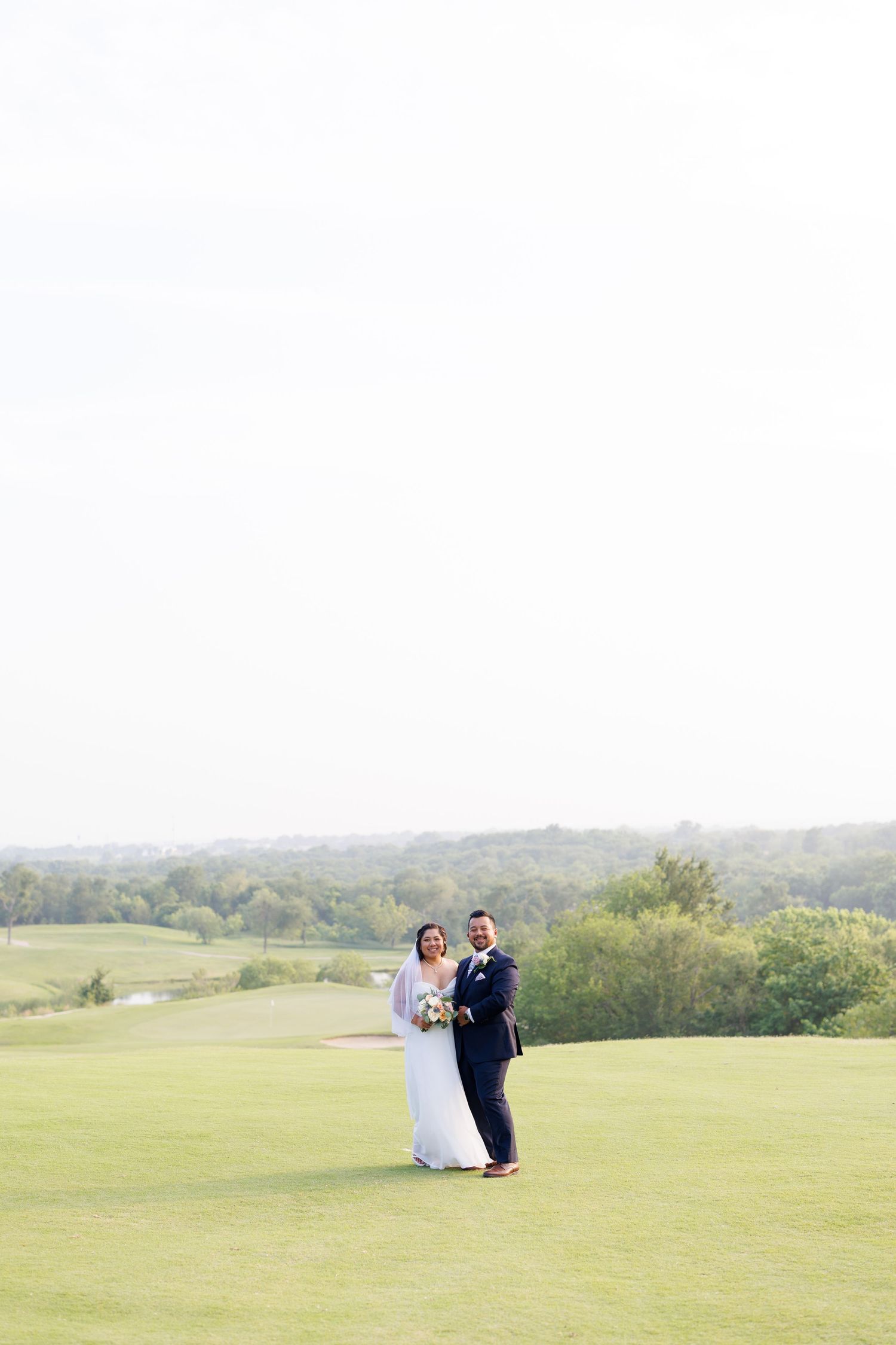 Newlyweds standing together on a lush golf course fairway with a scenic landscape backdrop.