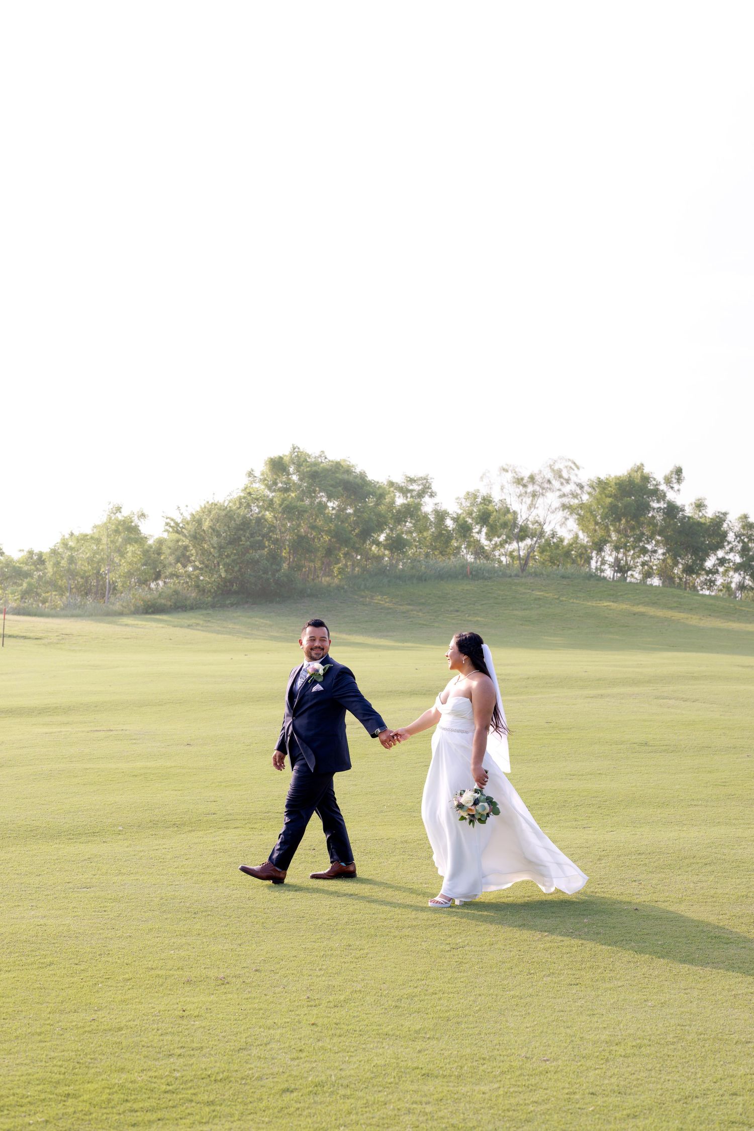 Bride and groom walk hand-in-hand across a lush green golf course on their wedding day.