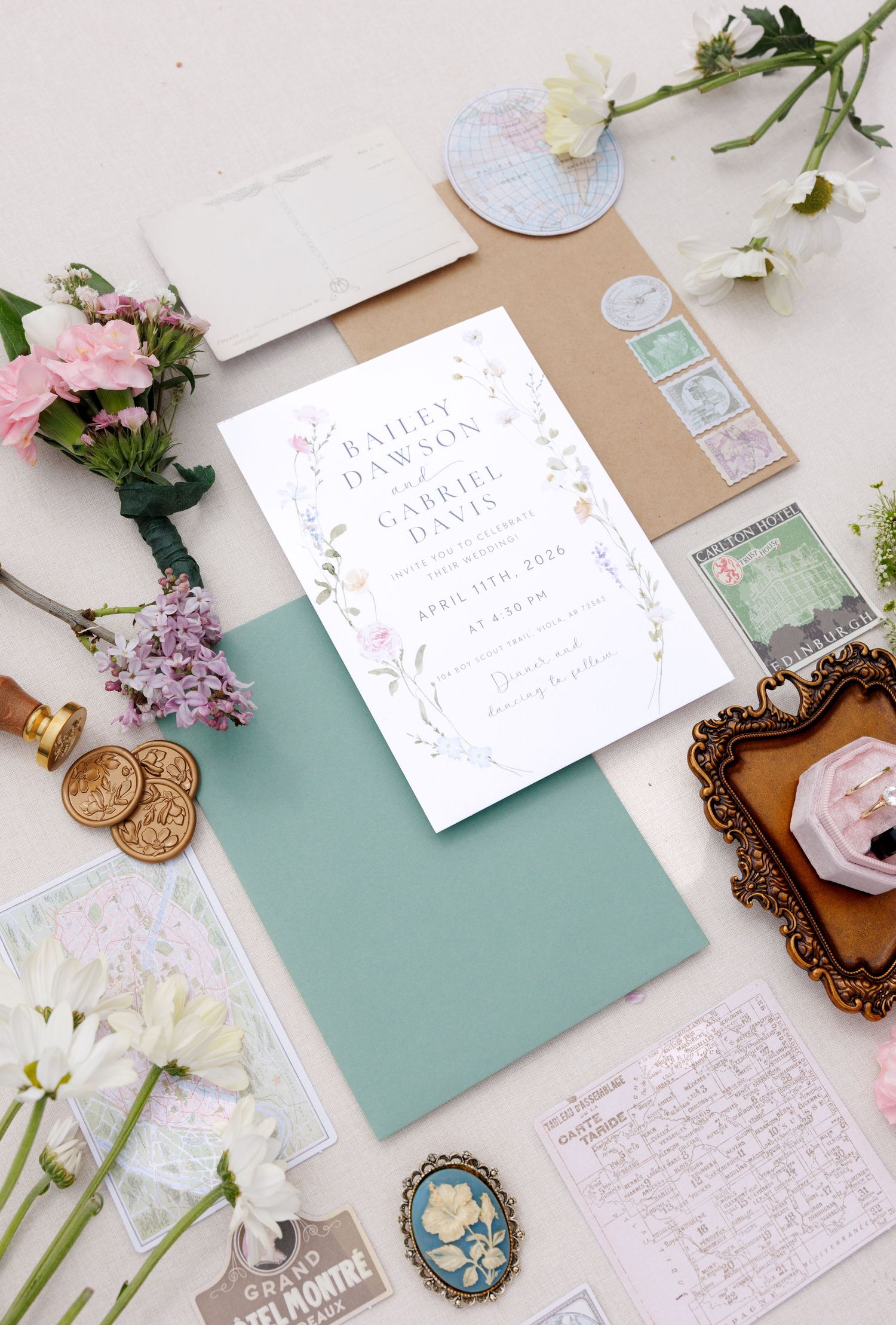 Elegant floral wedding invitation suite with sage green envelope, kraft paper, wax seals, stamps, and fresh flowers on white background.